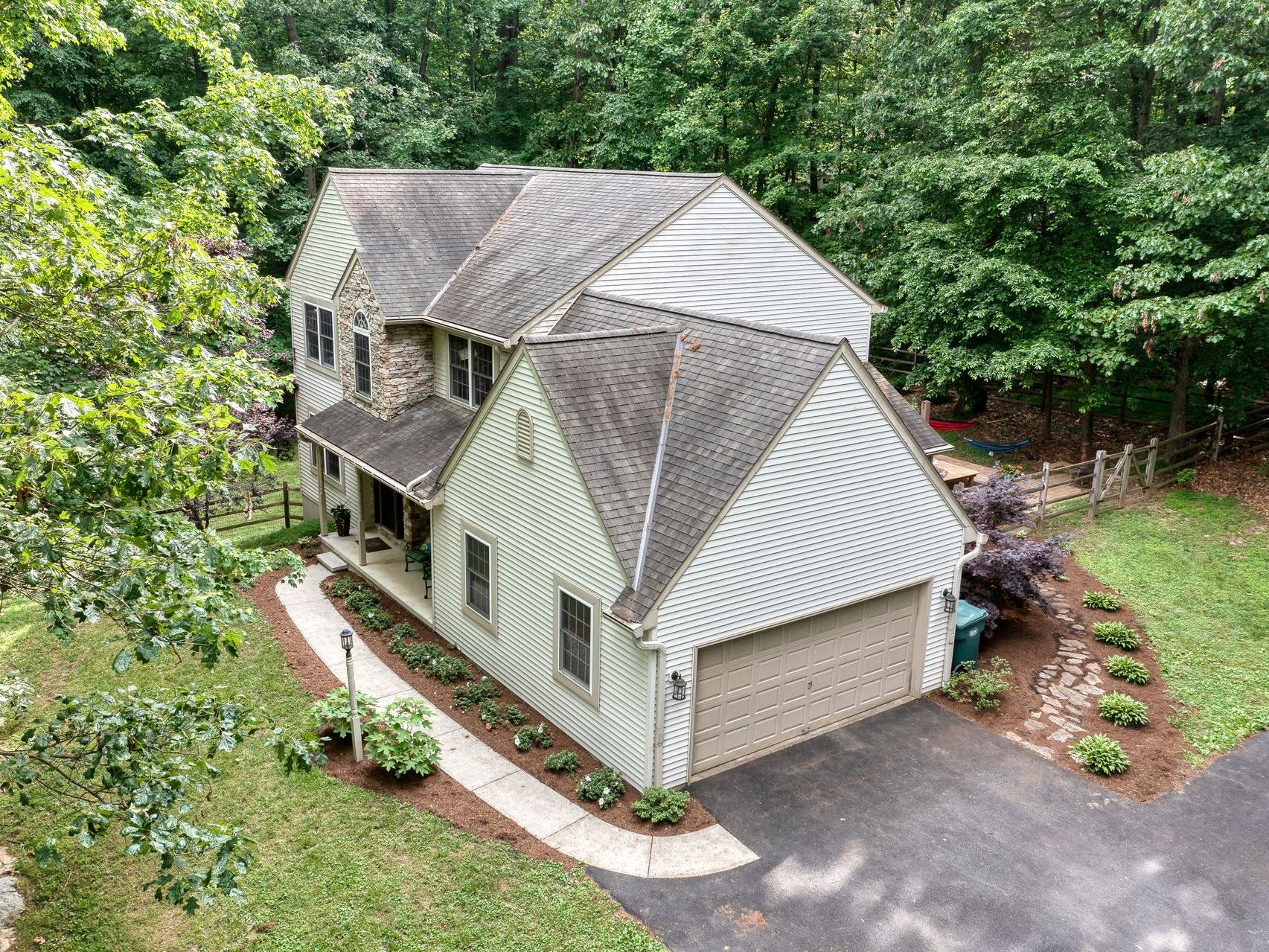Two-story house with tan siding, stone facade, and brown roof surrounded by trees and a driveway.
