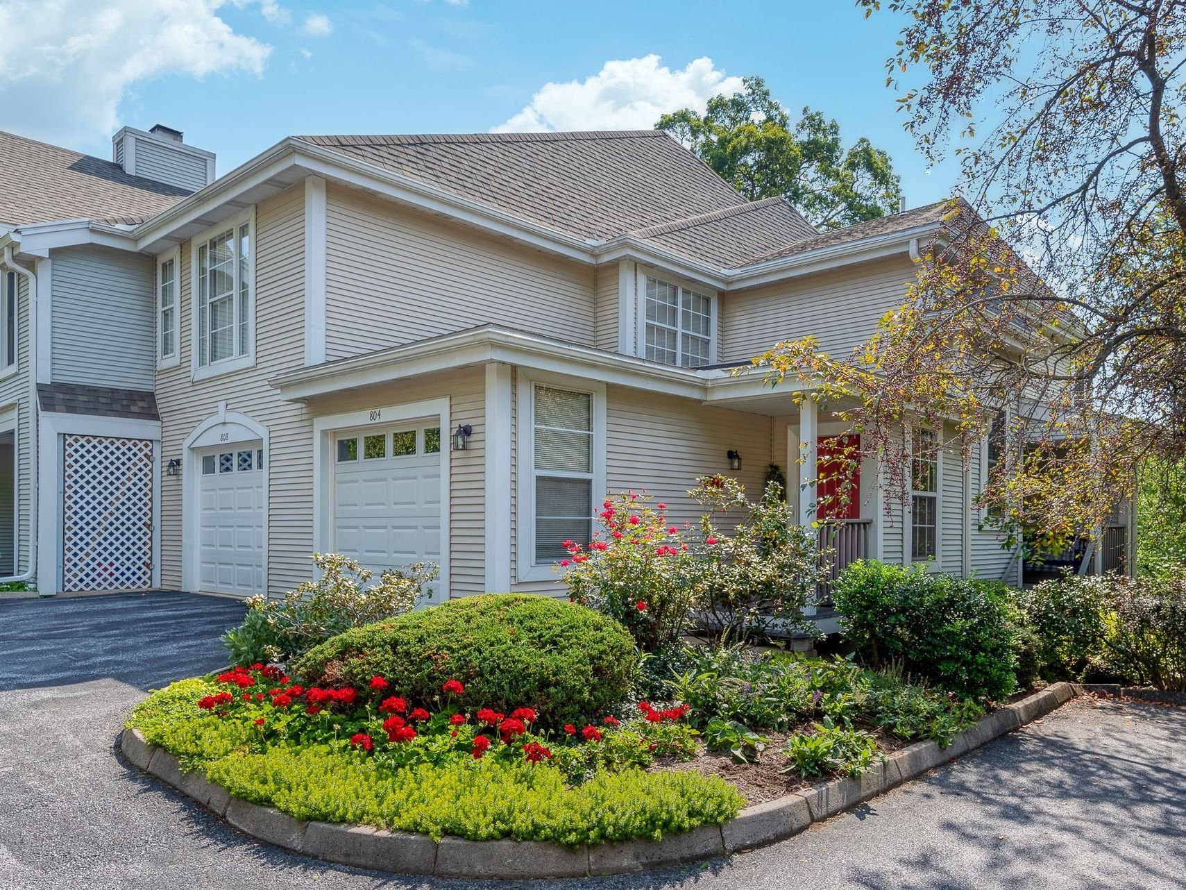 Beige two-story house with a garden, white garage doors, and a red front door on a sunny day.
