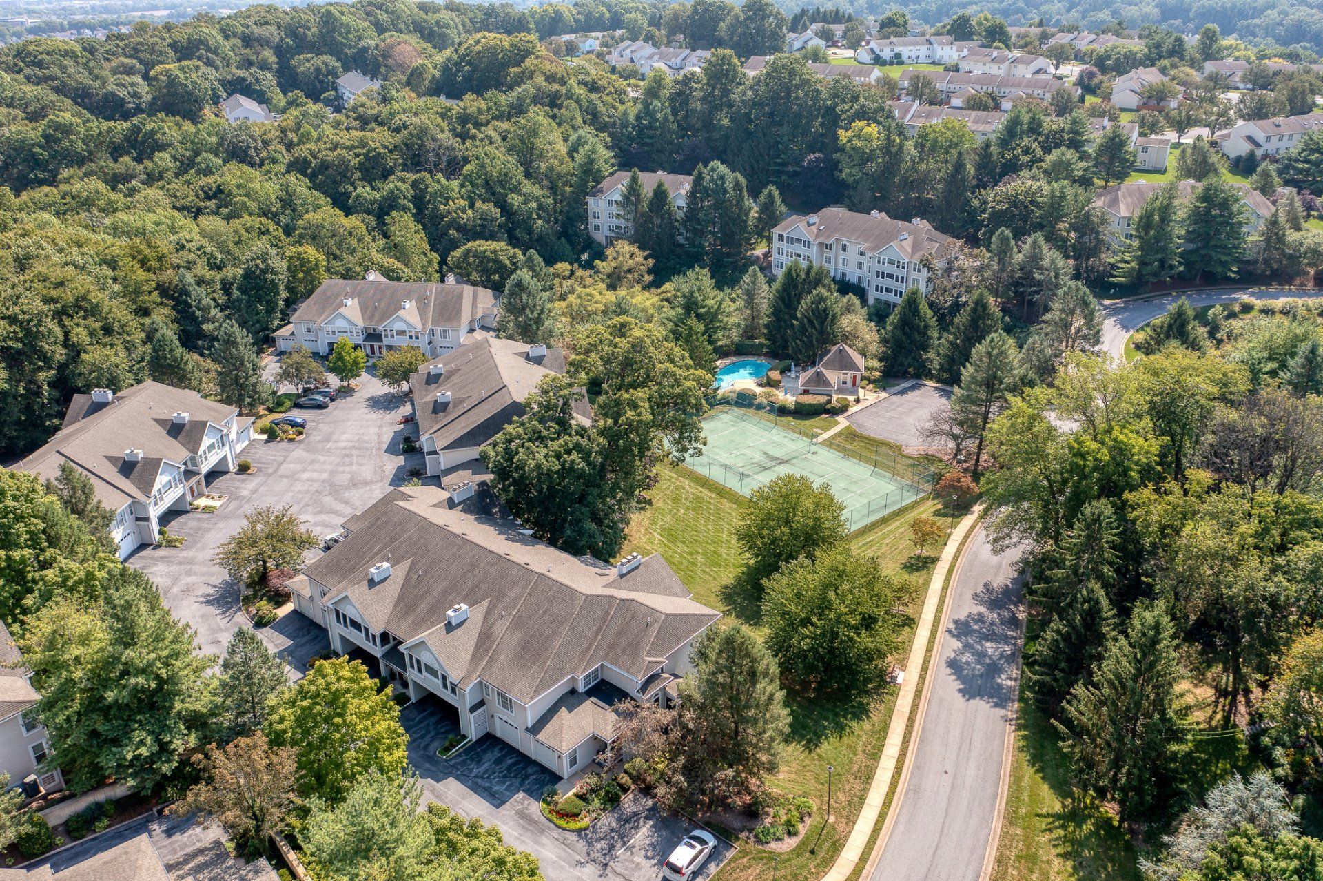 Aerial view of a residential area with buildings, trees, a pool, and tennis courts on a sunny day.