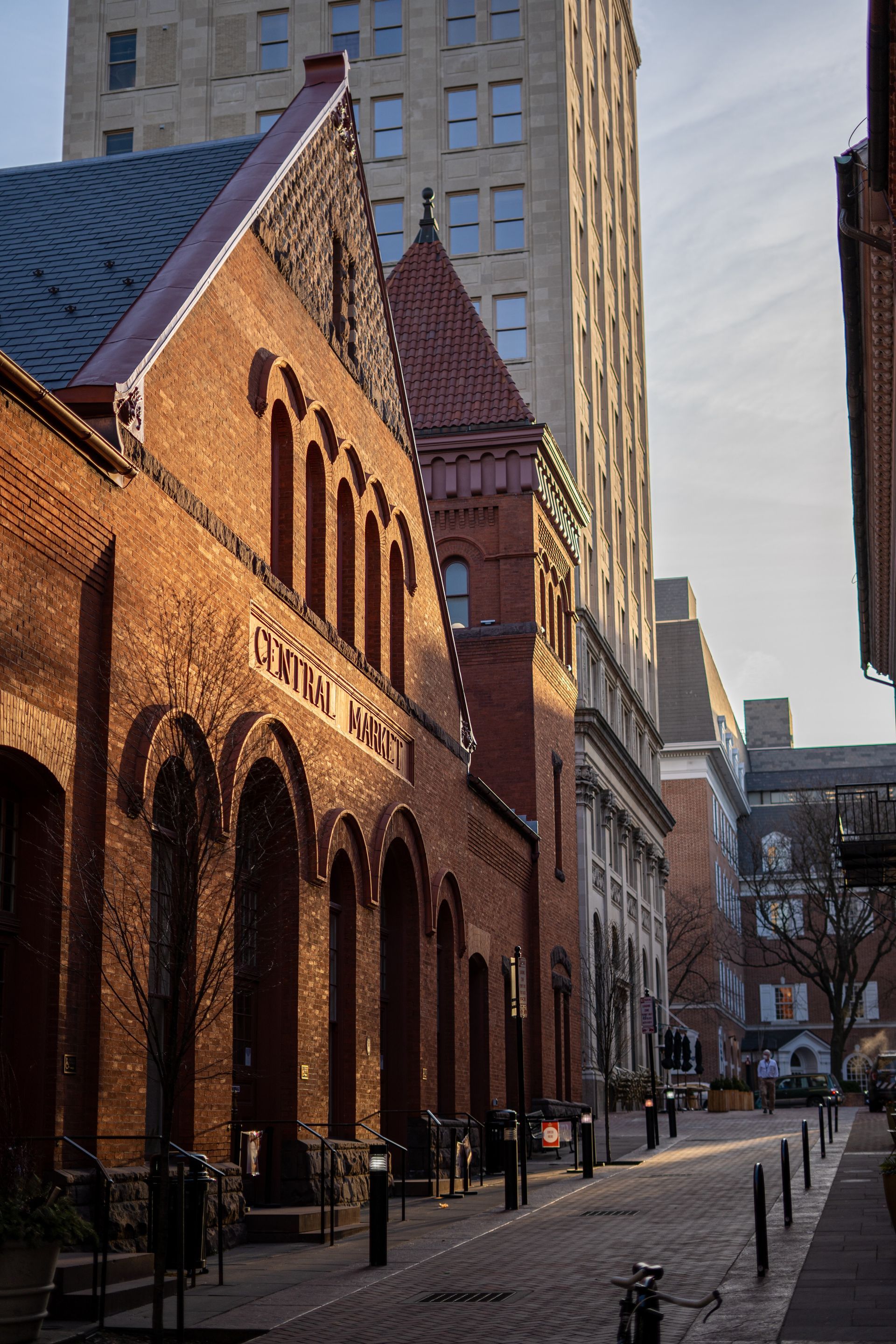 Brick buildings line a cobblestone street, bathed in sunlight. A tall building is in the background.