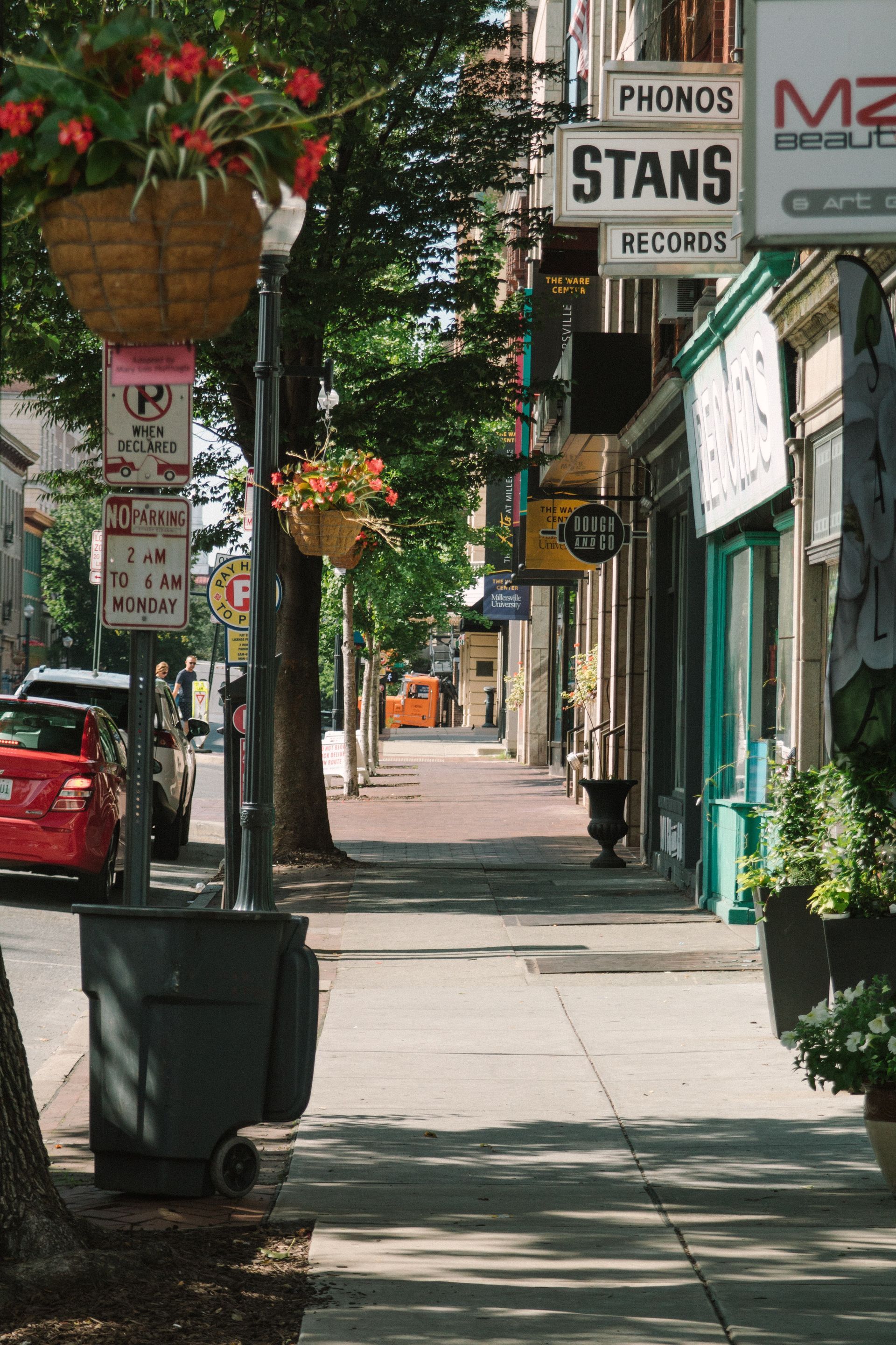 Sidewalk lined with shops, street signs, hanging baskets of flowers, and parked cars.