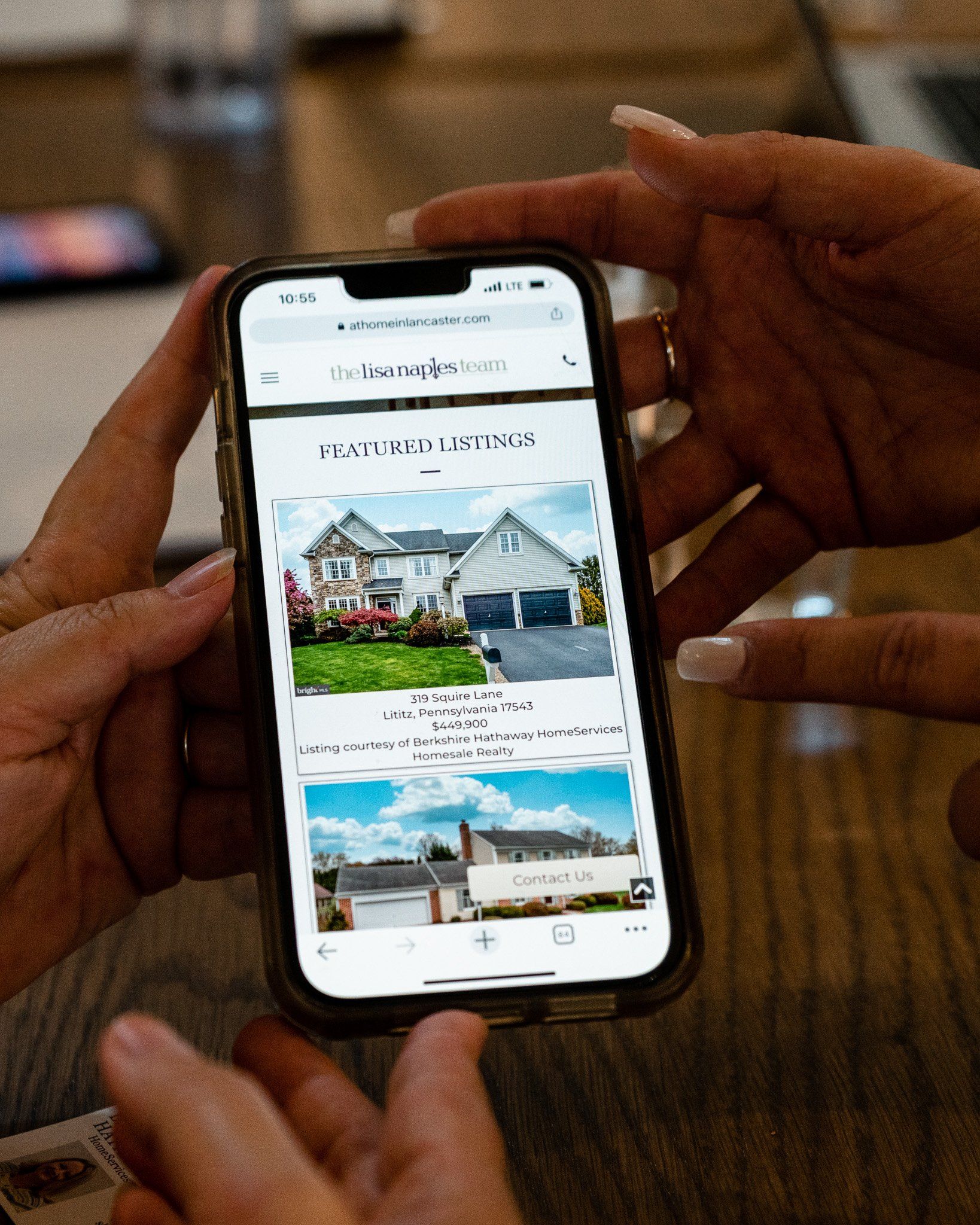 Hands holding a smartphone displaying a real estate listing with two house images, in a light setting.