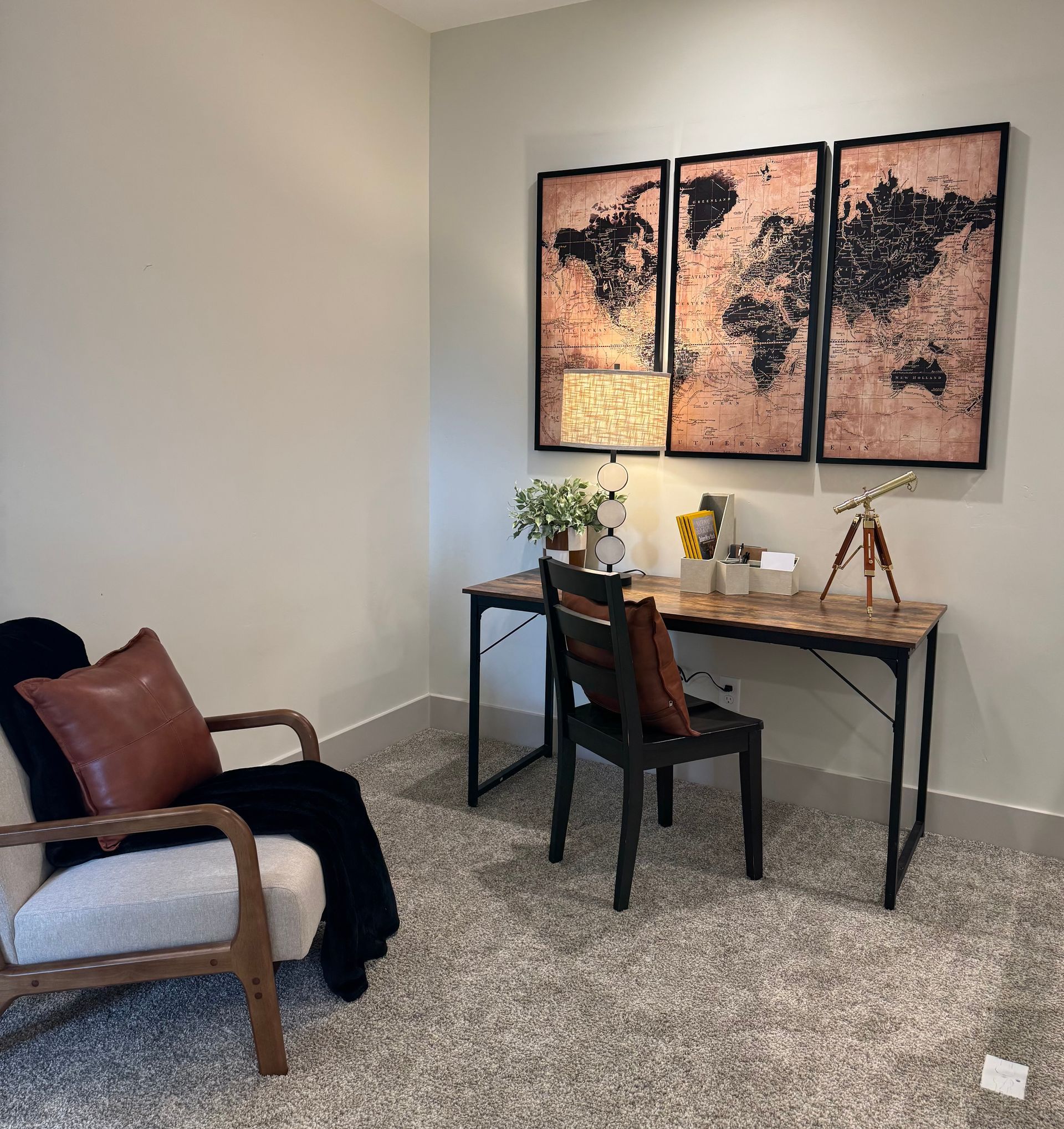 Office corner: desk with map art, chair, lamp, small telescope, and armchair with a pillow.