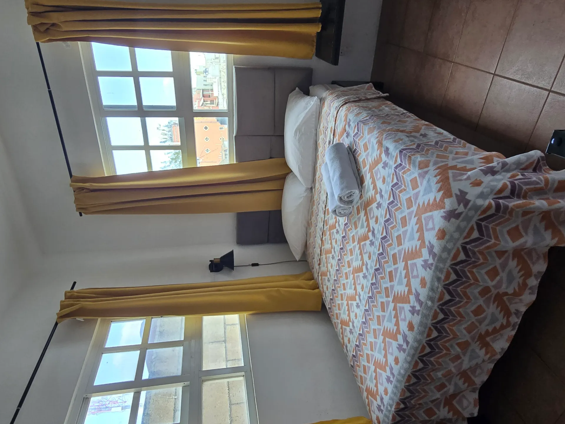 Habitación pequeña con una cama junto a dos ventanas, cortinas amarillas y suelo de baldosas.