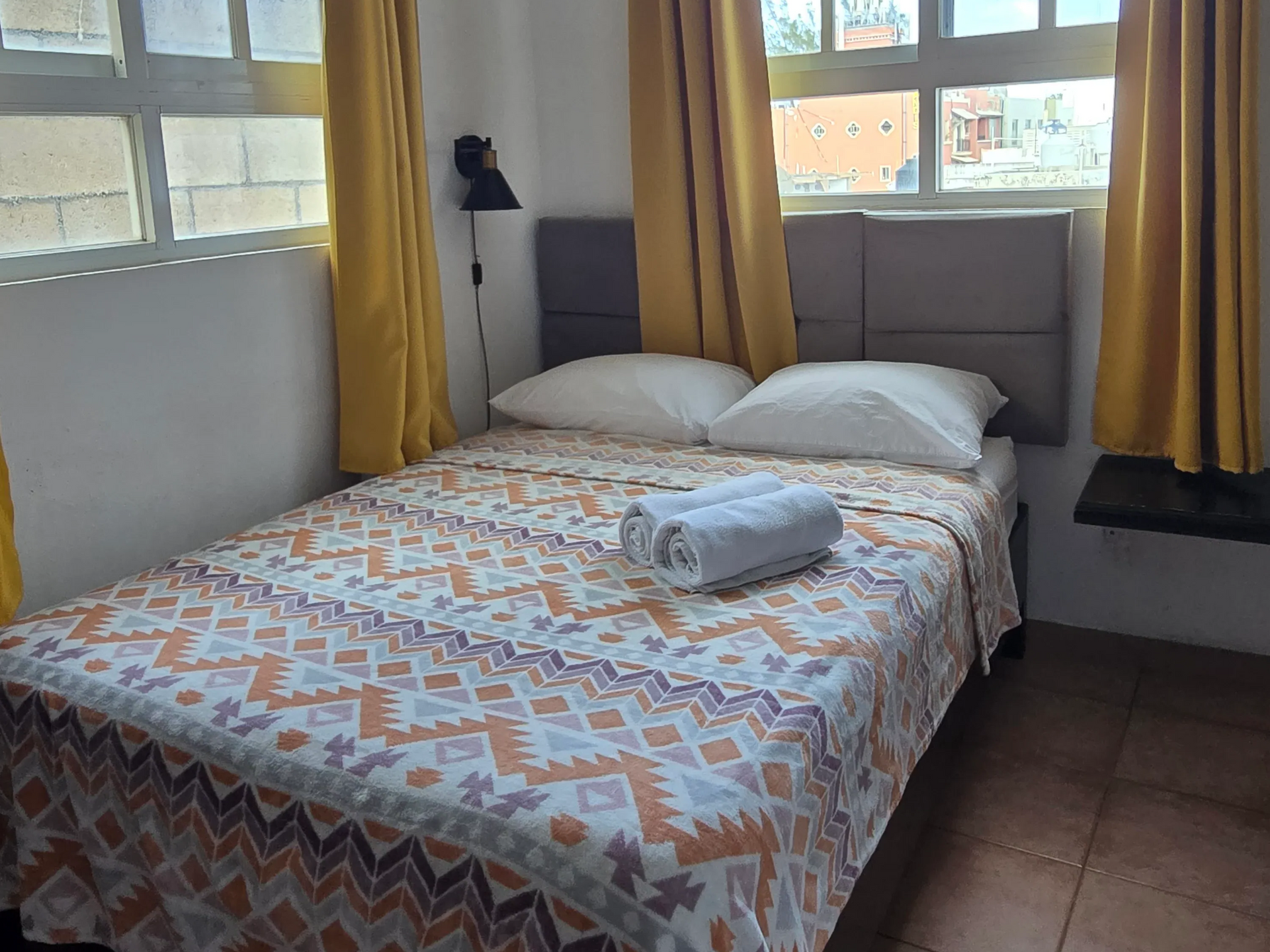 Habitación pequeña con cama hecha, toalla doblada, cortinas amarillas y ventanas que dejan entrar la luz del día.