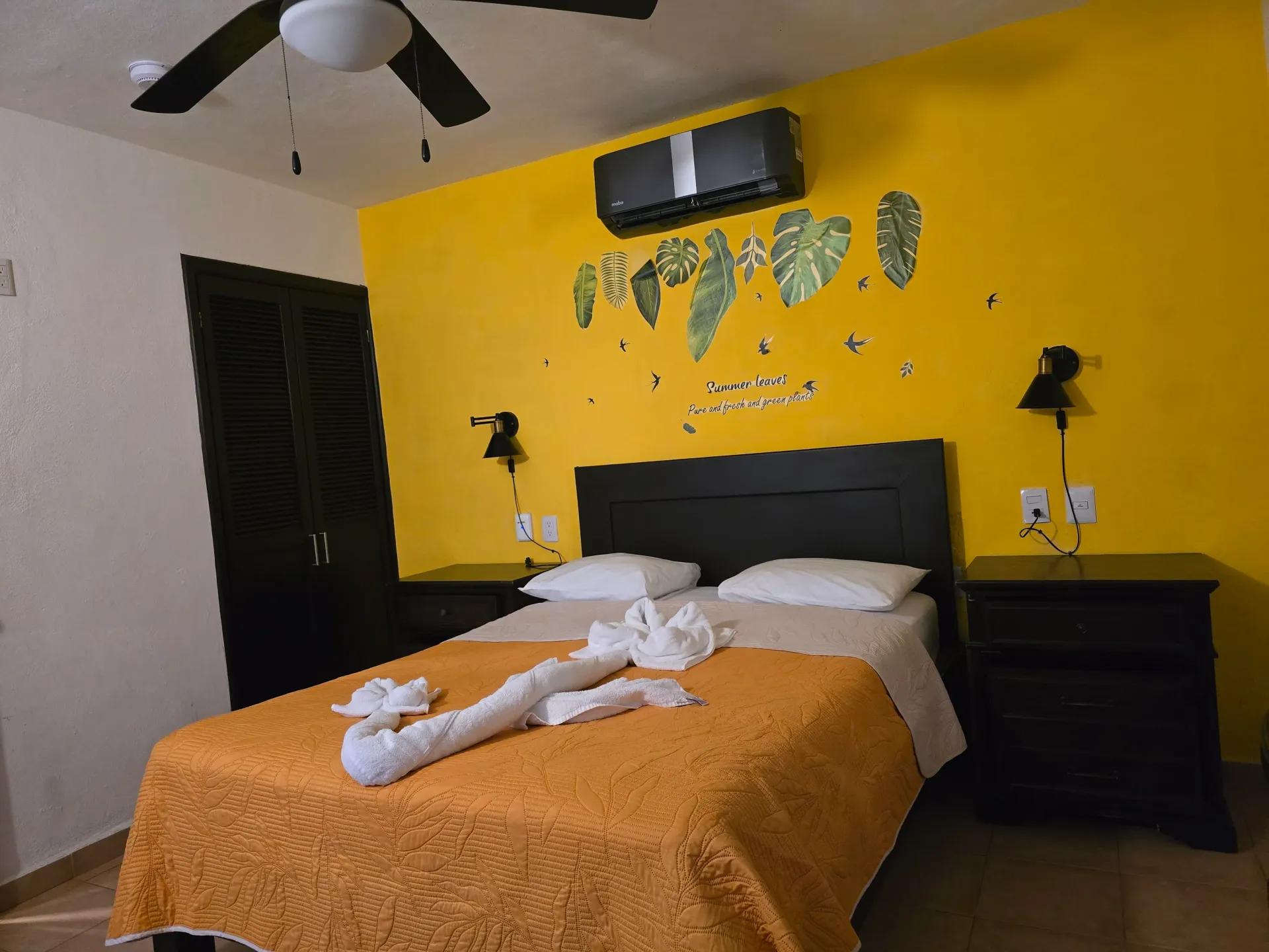 Dormitorio con pared decorativa amarilla, cama con manta naranja y un animal de toalla sobre la cama.