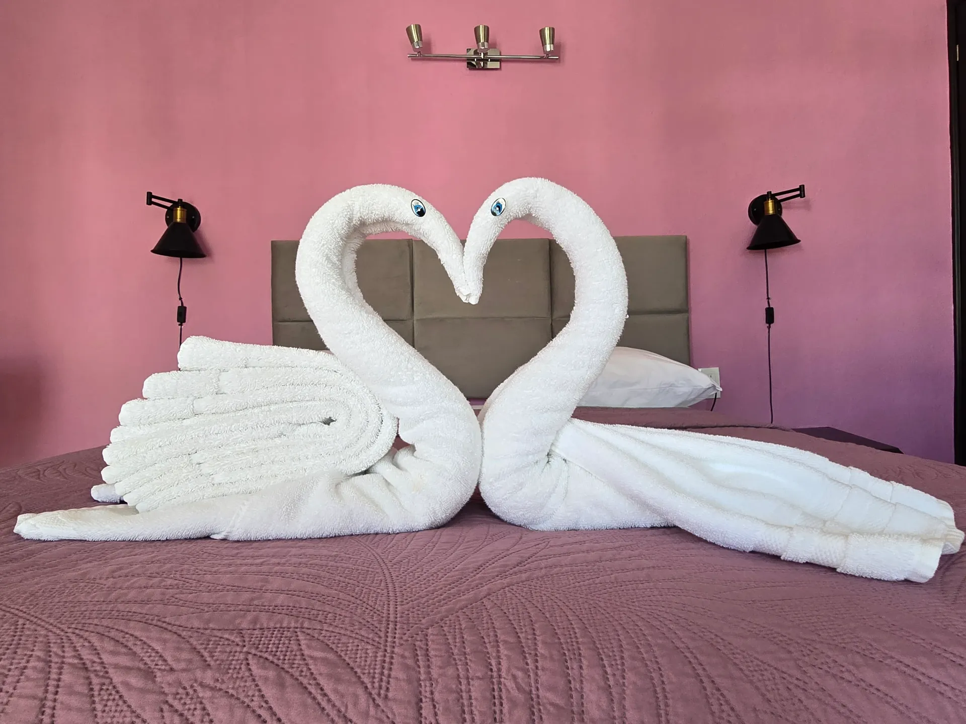 Dos esculturas de toallas con forma de cisne blanco forman un corazón sobre una cama de hotel rosa.