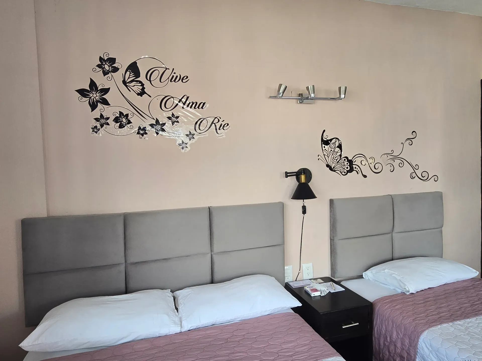 Dos camas con cabeceros grises en un dormitorio con vinilos decorativos florales en pared y una pequeña lámpara.