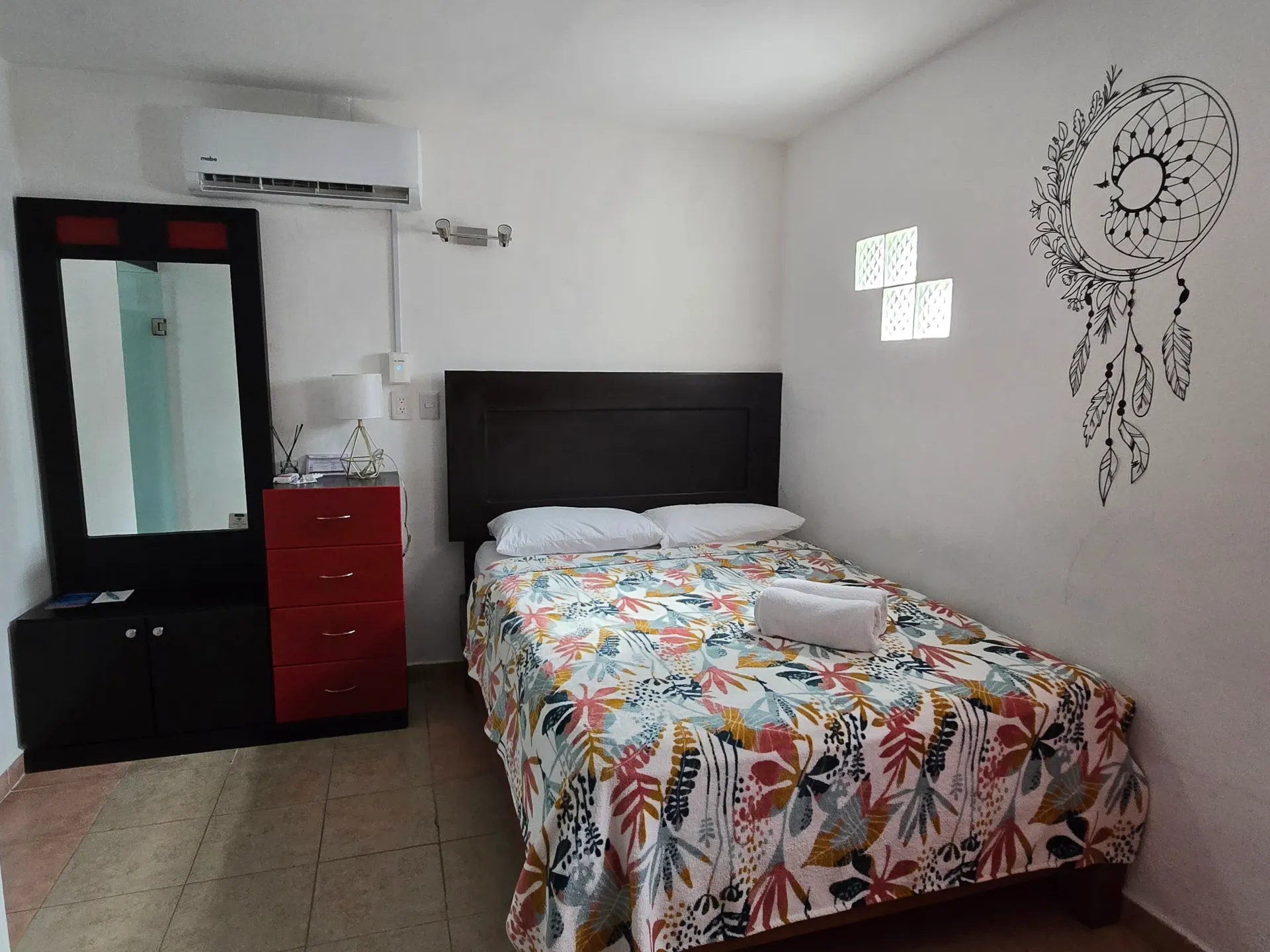 Habitación pequeña con cama, cómoda roja, espejo, aire acondicionado y un atrapasueños decorativo en la pared.