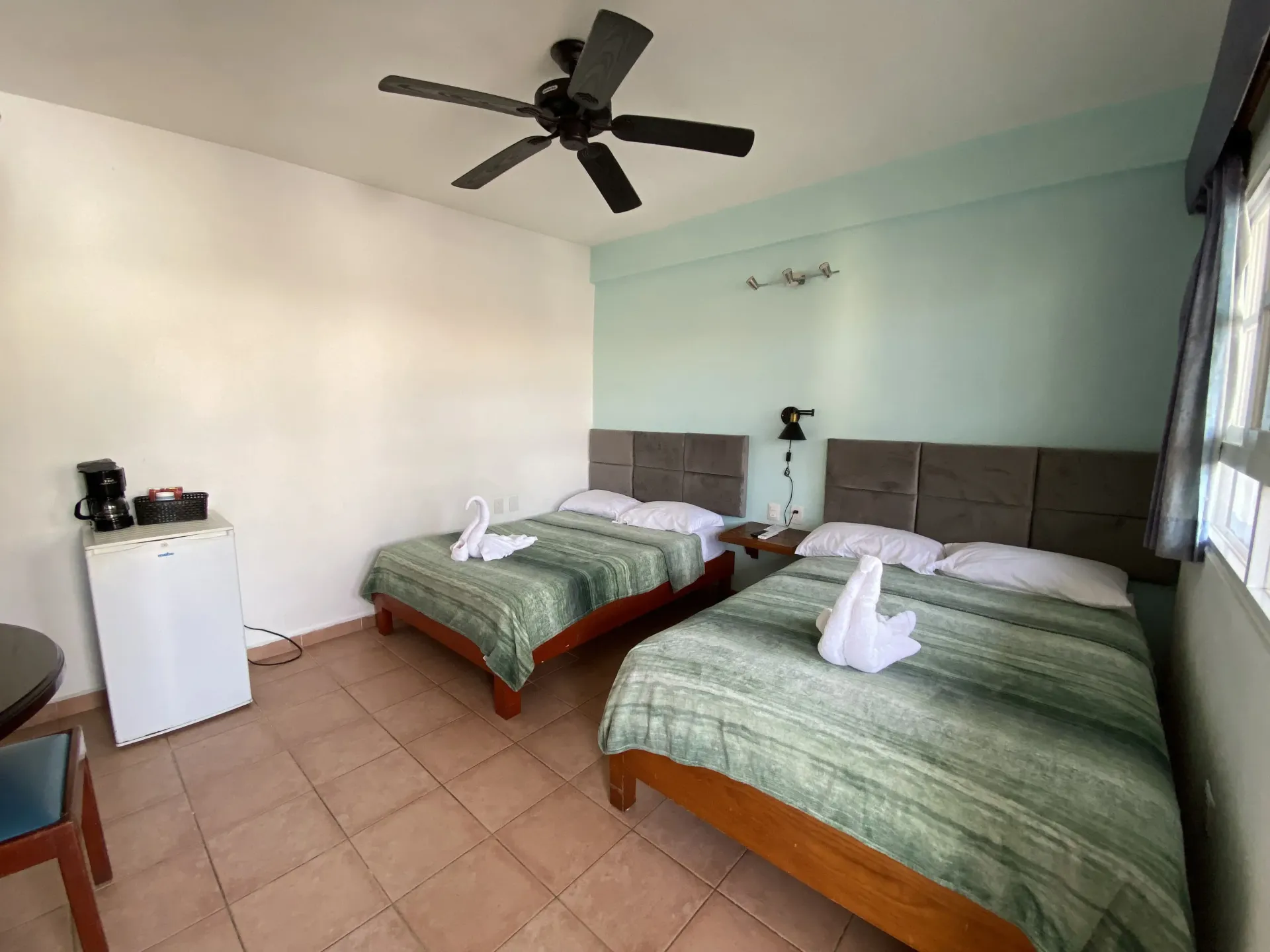 Dos camas individuales bien hechas en una luminosa habitación de hotel con ventilador de techo y suelo de baldosas.
