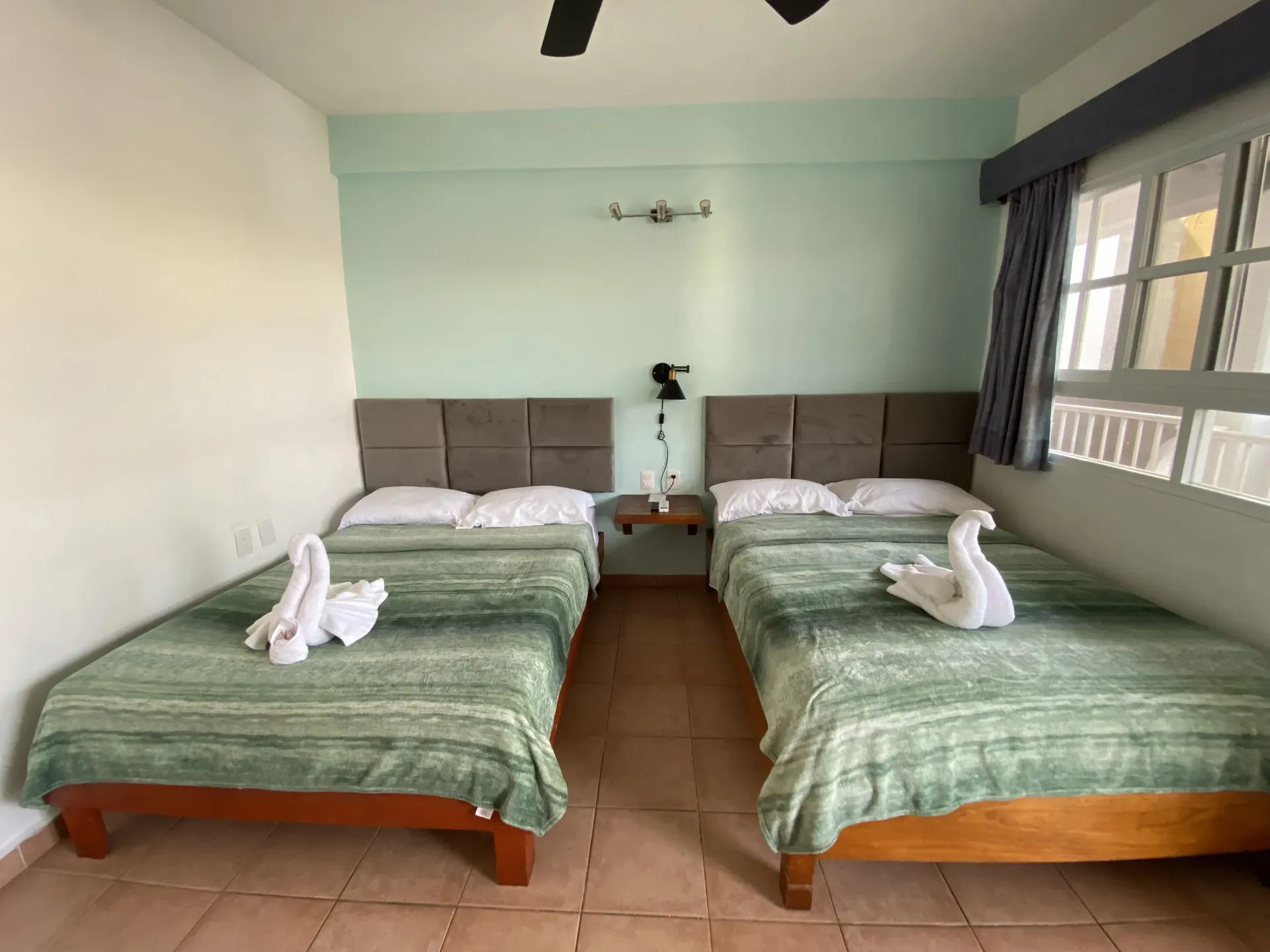 Dos camas individuales en una habitación luminosa con edredones verdes, cabeceros grises y cisnes hechos con toallas .