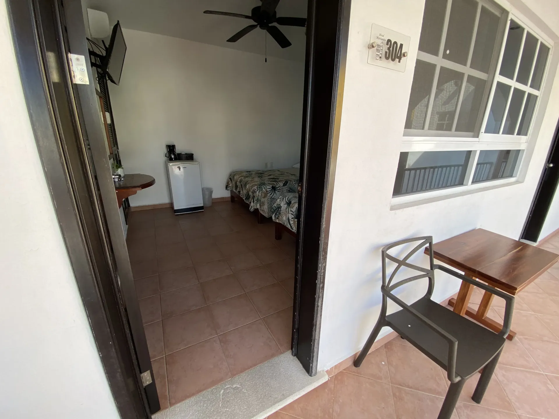 Entrada abierta a una habitación con piso de baldosas, cama, ventilador de techo y una silla pequeña en un patio.