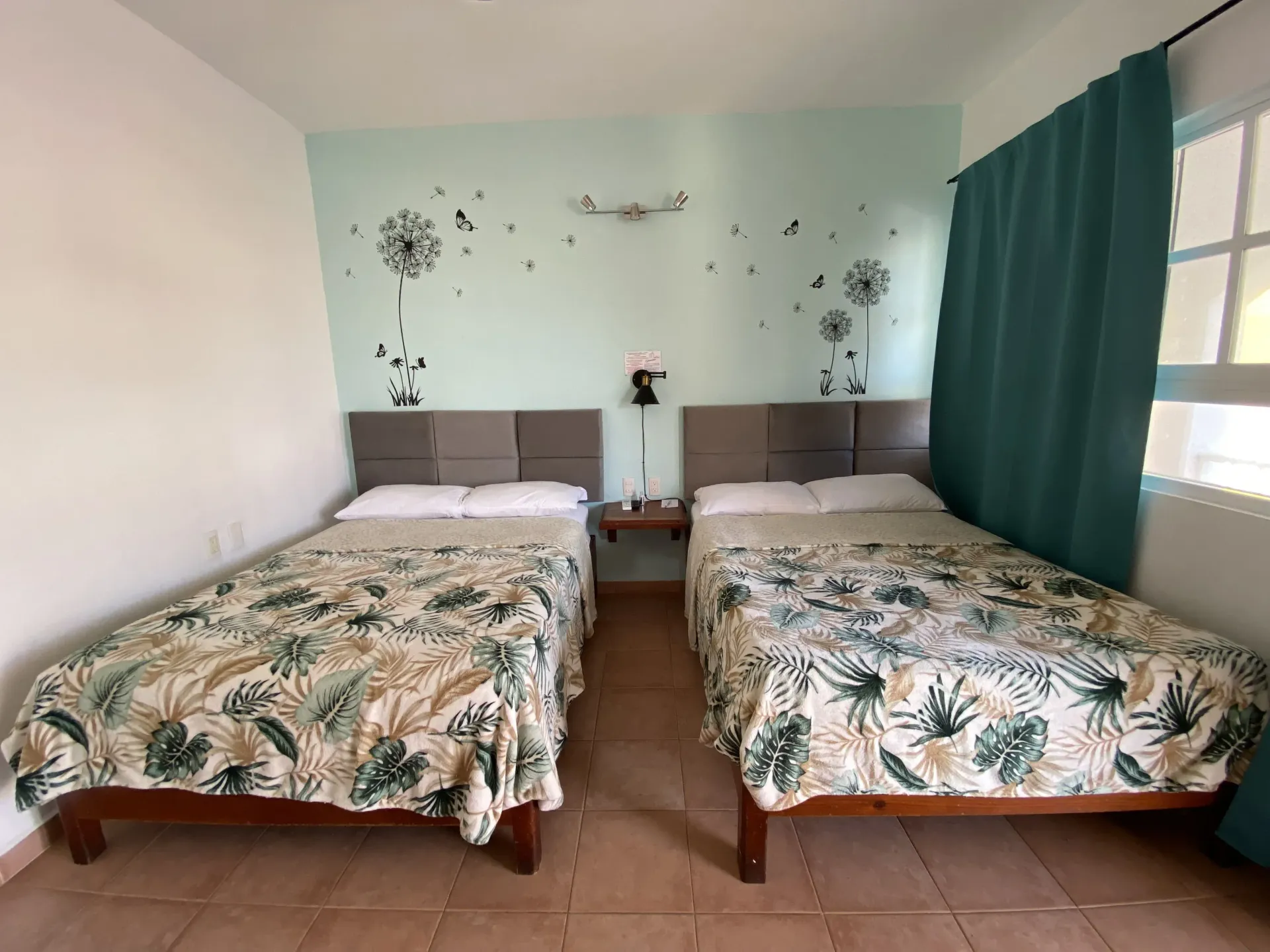 Habitación con dos camas individuales, colchas con estampado de hojas verdes y una cortina color turquesa junto a la ventana.