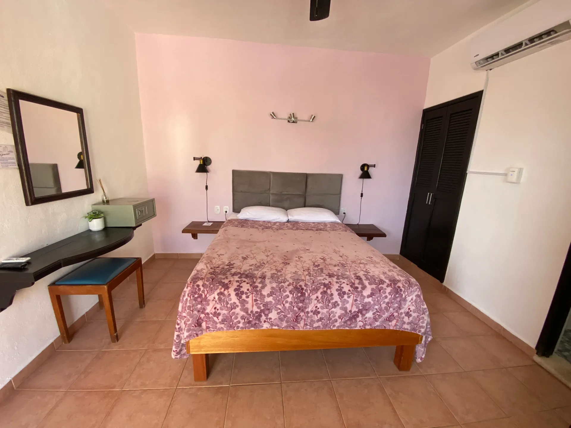 Dormitorio con cama, escritorio, banco, espejo de pared y puertas negras sobre suelo de baldosas.