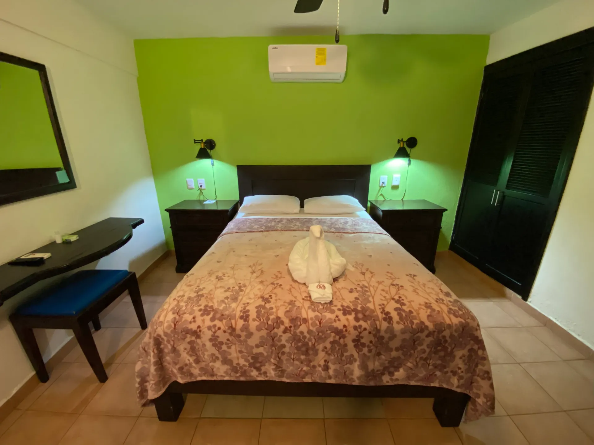 Dormitorio con paredes verdes, una cama bien hecha, lámparas de noche y un escritorio junto a la ventana.