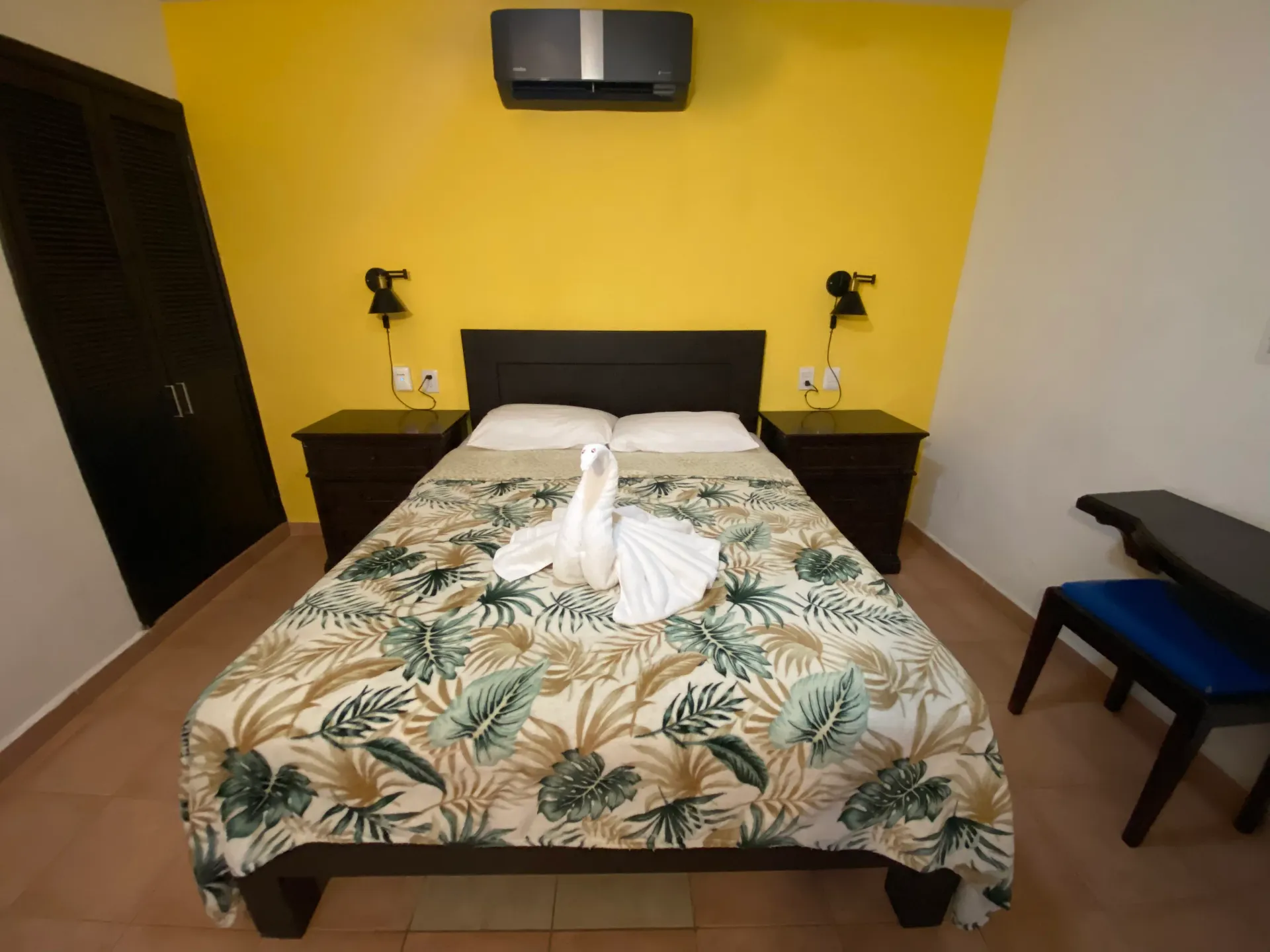 Habitación con una cama , una pared decorada en amarillo, una t.v. montada en la pared y una silla junto a la ventana.