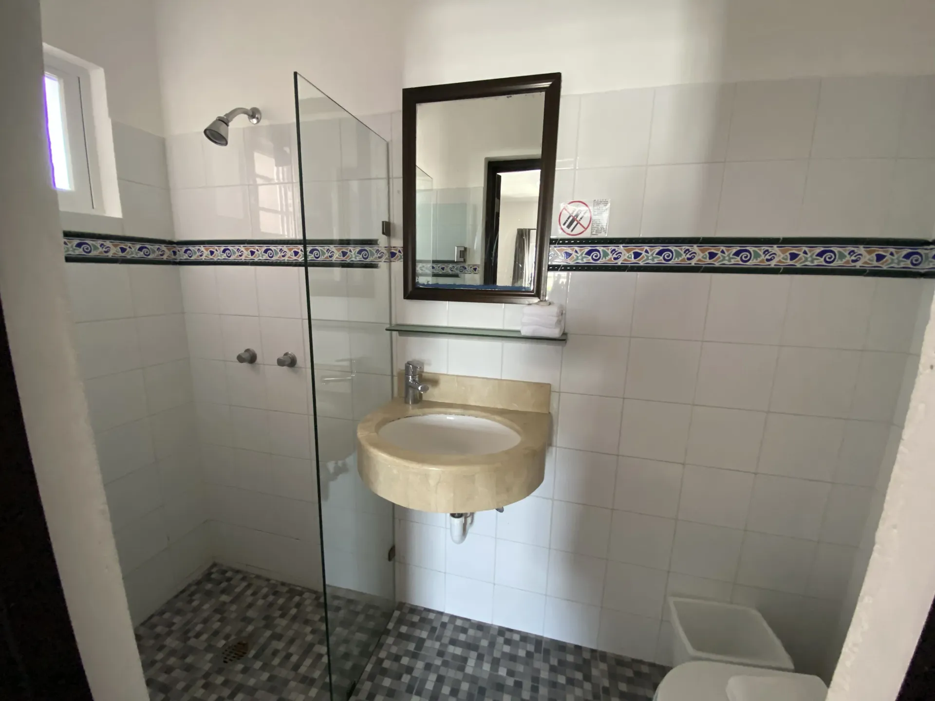 Pequeño baño alicatado con ducha de cristal, lavabo mural, espejo y portarrollos de papel higiénico.
