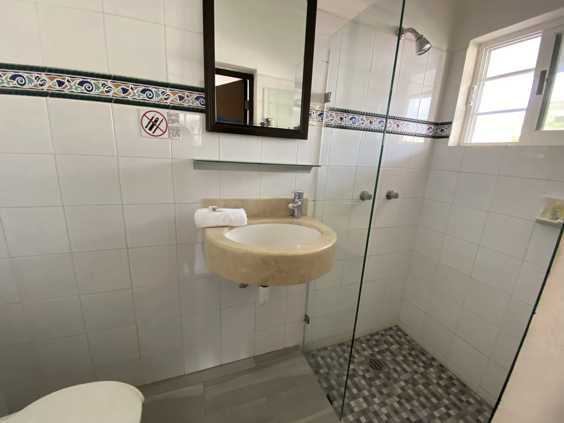 Baño alicatado con lavabo mural, espejo, portarrollos de papel higiénico y cabina de ducha de cristal con suelo de guijarros.