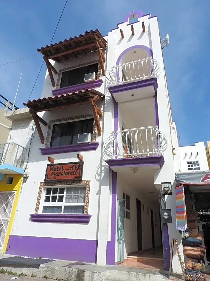 Edificio de varias plantas de color morado y blanco, con balcones y una entrada estrecha, situado en una calle soleada.