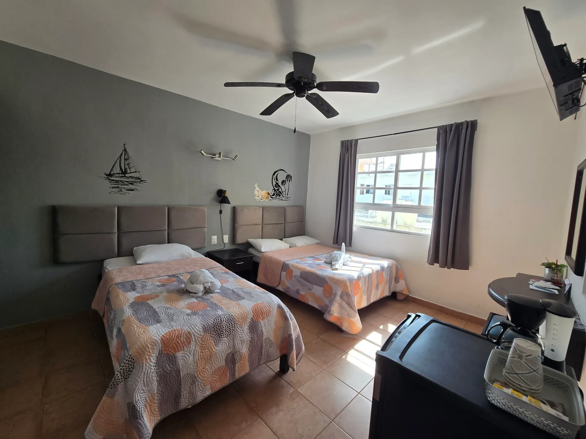 Dos camas individuales en una sencilla habitación de hotel con ventilador de techo, ventana y suelo de baldosas.