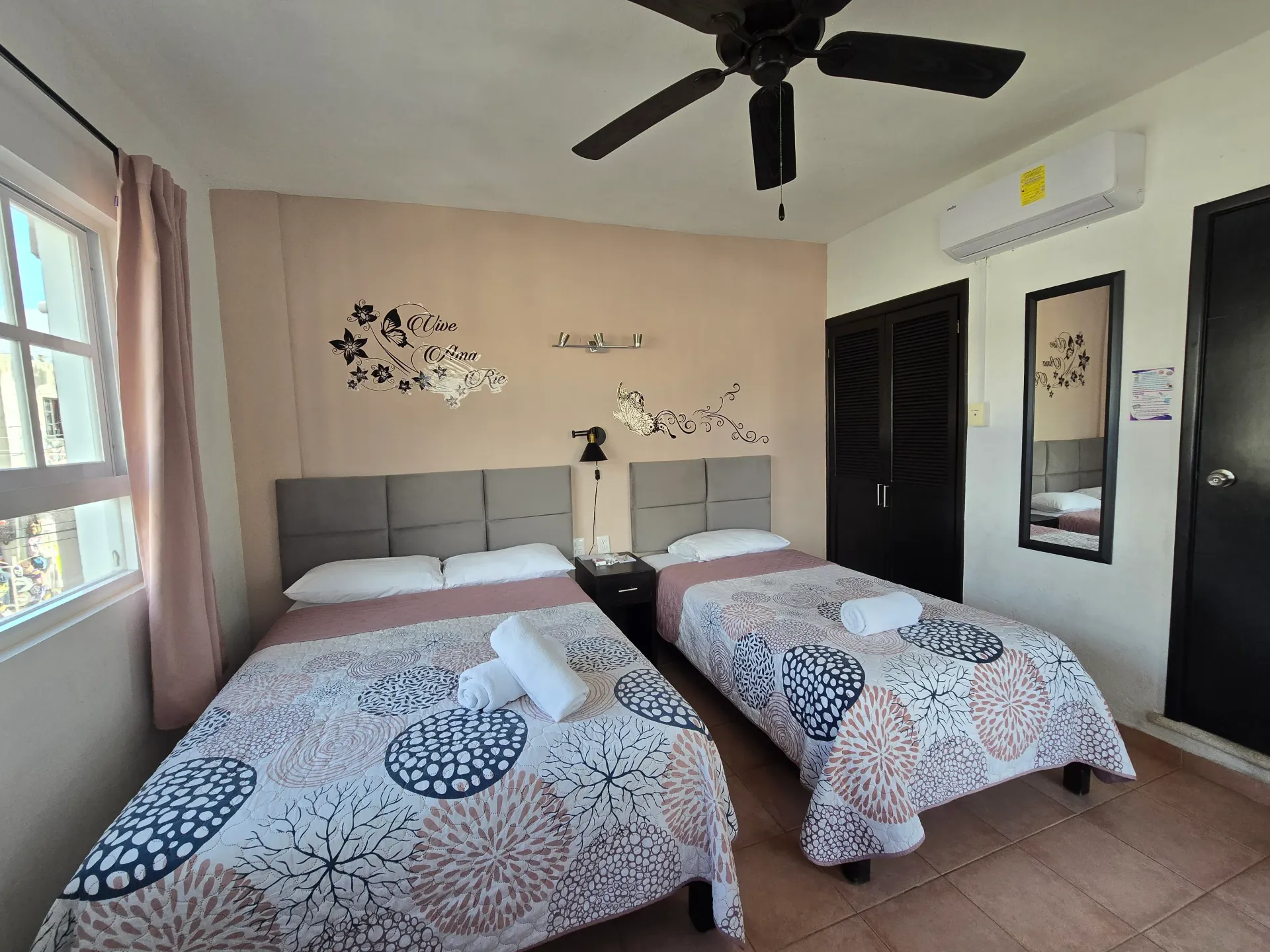 Habitación con dos camas, ventilador de techo, ventana grande y paredes de color beige con cuadros.