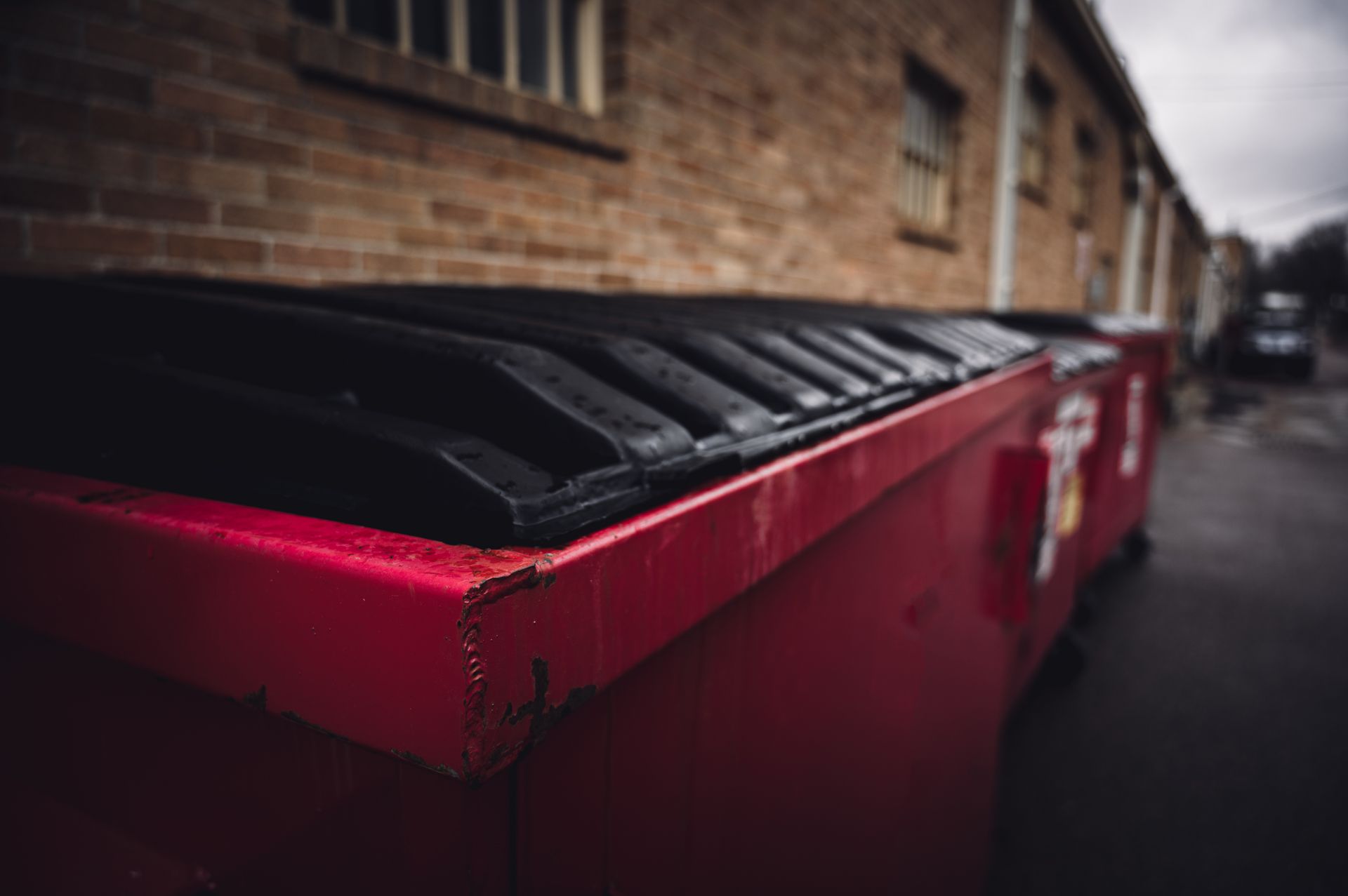 Long red metal dumpster with black lids positioned along a brick building in an alleyway.