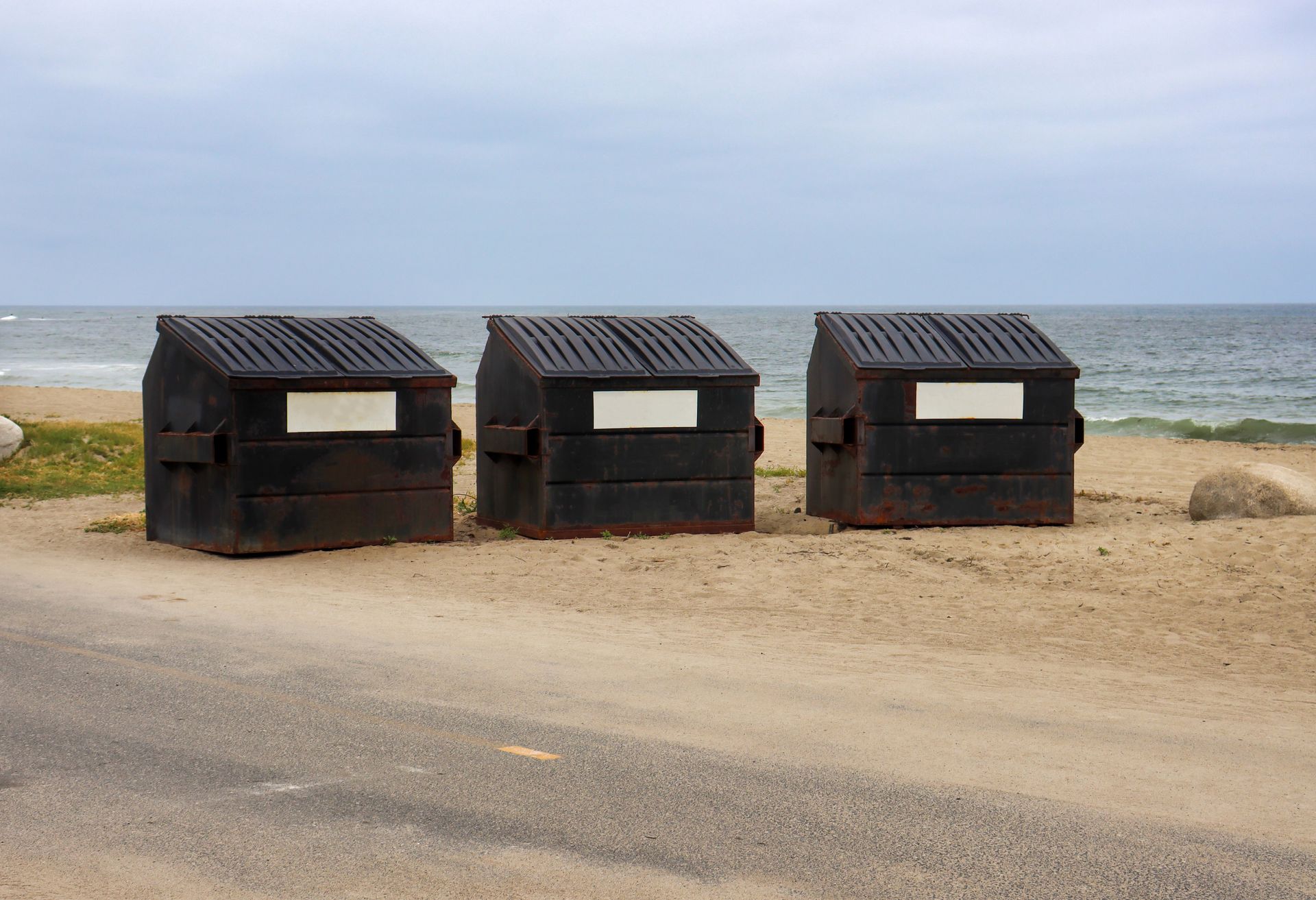 Three black dumpsters on sand beside a road near the beach.
