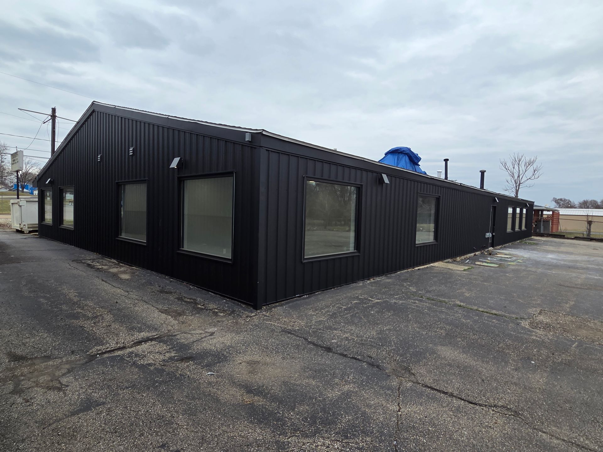 Black corrugated-metal building with large windows on a paved lot under cloudy skies