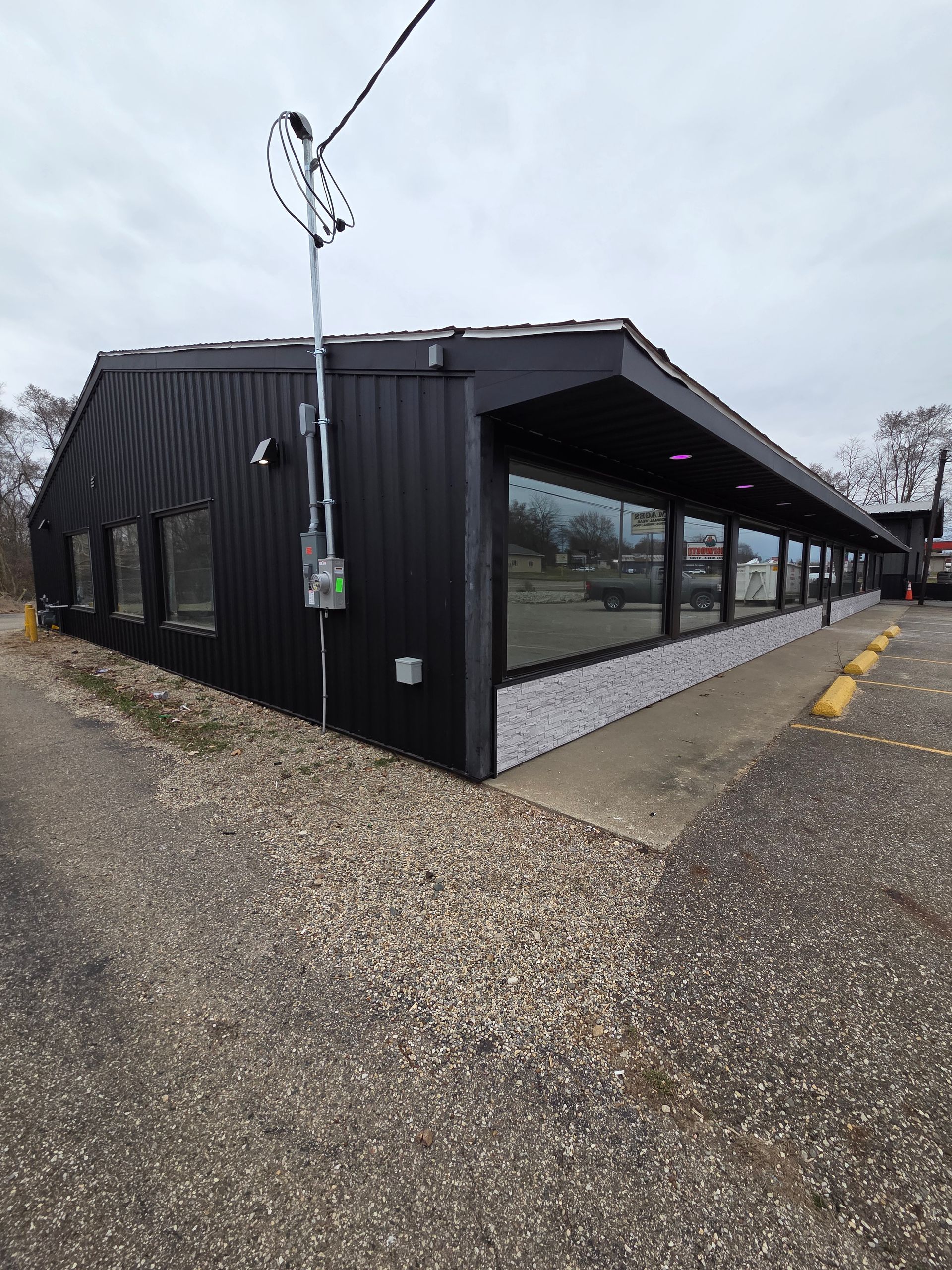 Black corrugated building with large windows on a gravel lot under a cloudy sky