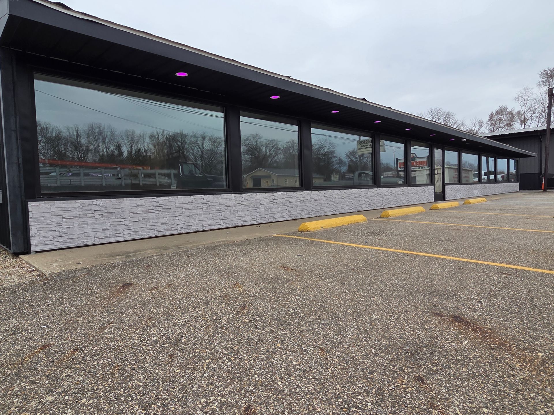 Empty parking lot beside a long, dark building with large windows under an overcast sky.