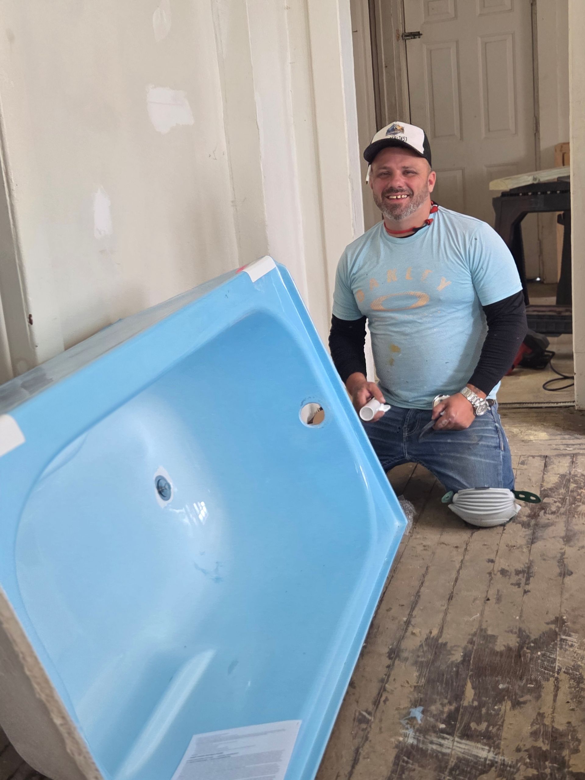Man kneeling beside a blue bathtub in a room under renovation, holding tools and smiling.