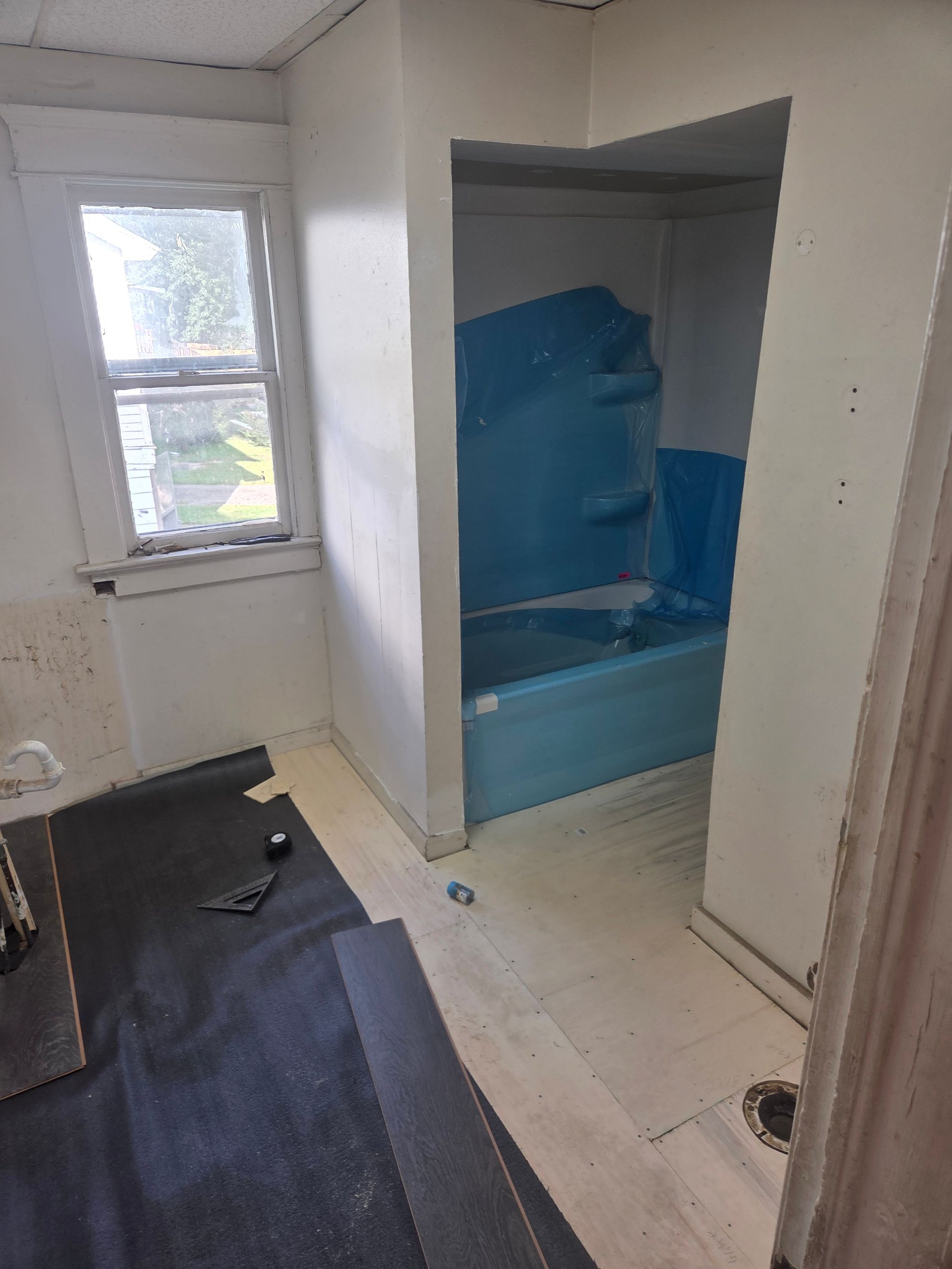 Small room with a window, black floor mat, and a blue bathtub in a tiled alcove.