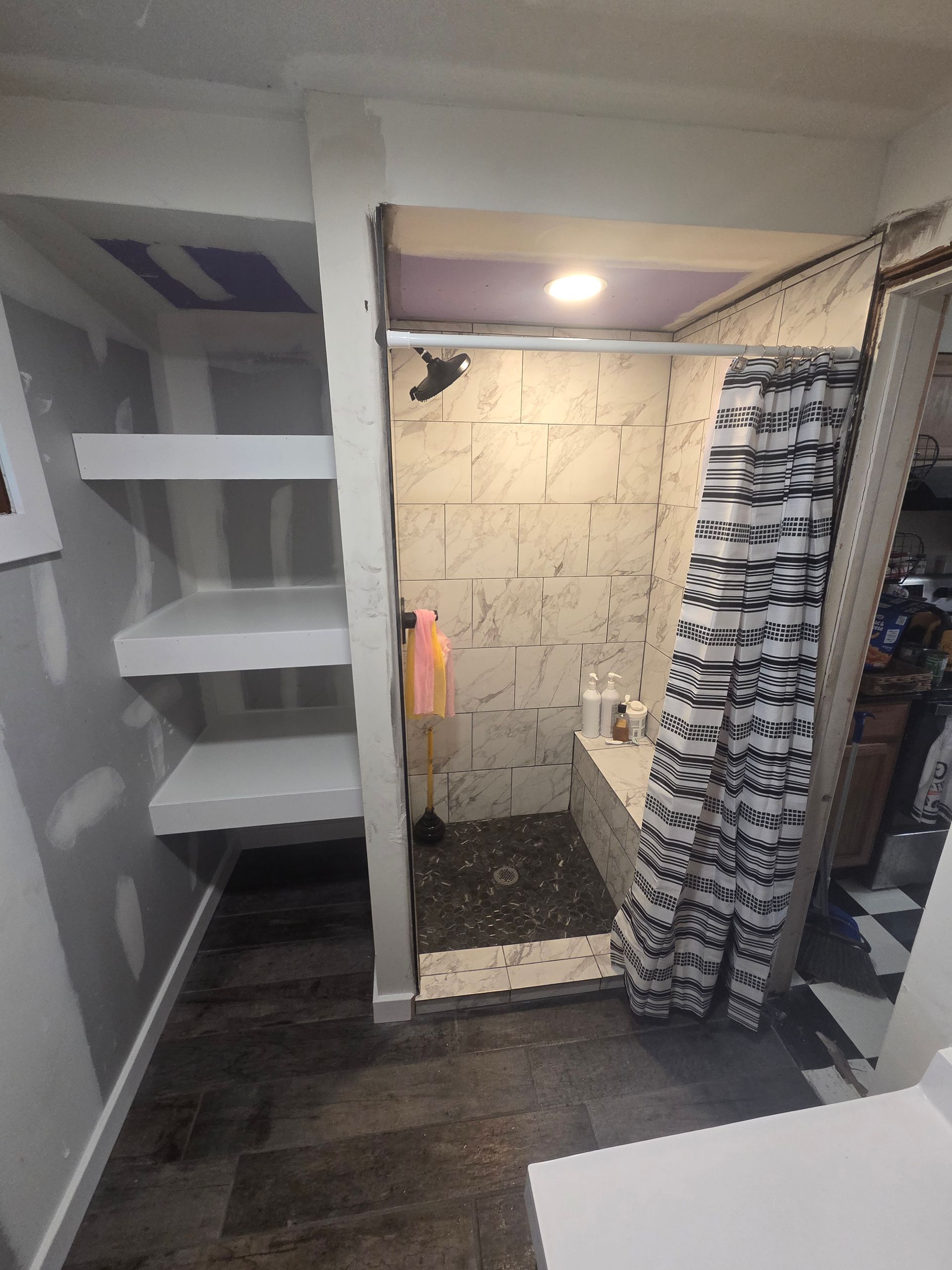 Small closet bathroom with shelves, tiled shower, and a striped curtain in a compact space.