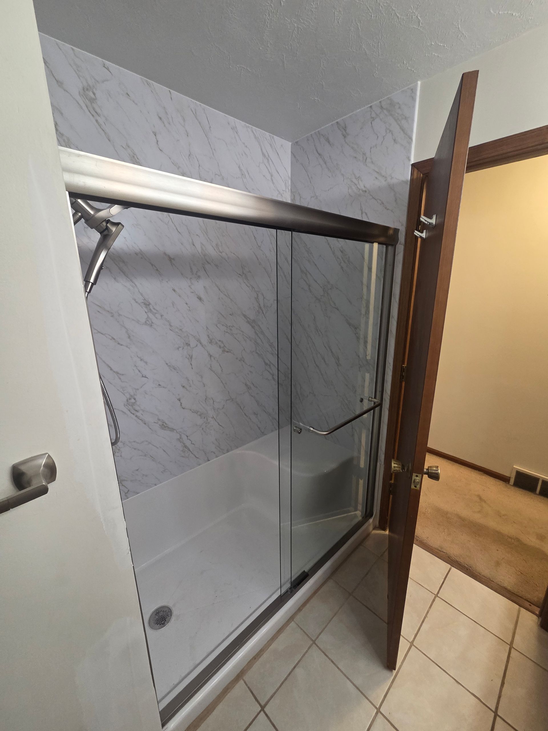 Bathroom shower stall with glass sliding door, marble-look tile, and open wooden door nearby