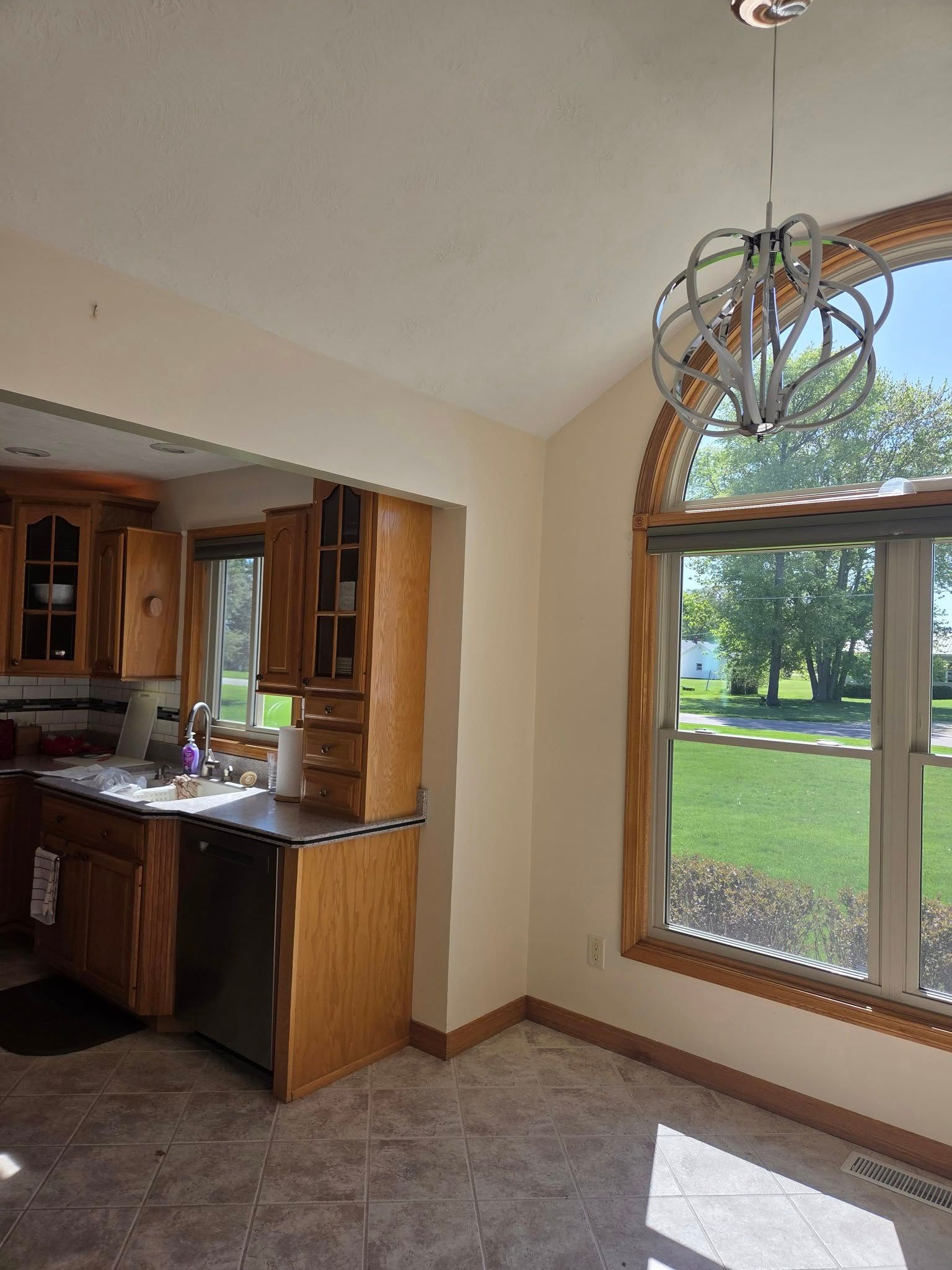 Empty dining area with large arched window, chandelier, and view of a green yard beyond.