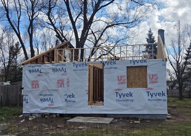 Partially built small house wrapped in Tyvek, with roof framing and plywood window openings under bare trees