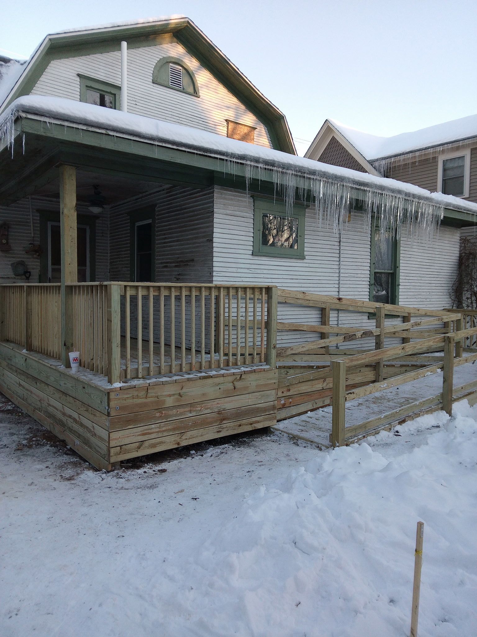 Snow-covered house with wooden porch and railing decorated with icicles, viewed in winter light