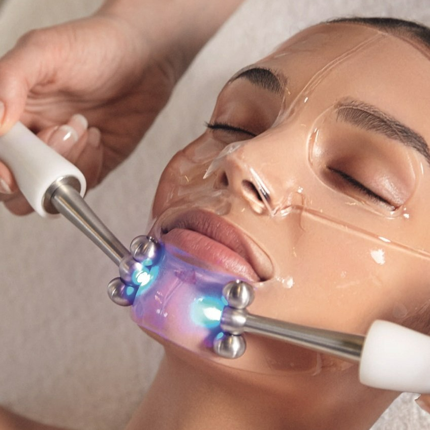 Person receiving facial treatment with a mask and massage tool, lit by blue light.