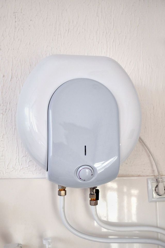 White electric water heater mounted on a wall with two pipes and a control knob.