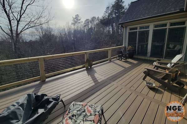 Wooden deck with cable railing overlooking a forested area. Chairs, sunlight, and a sunroom are visible.
