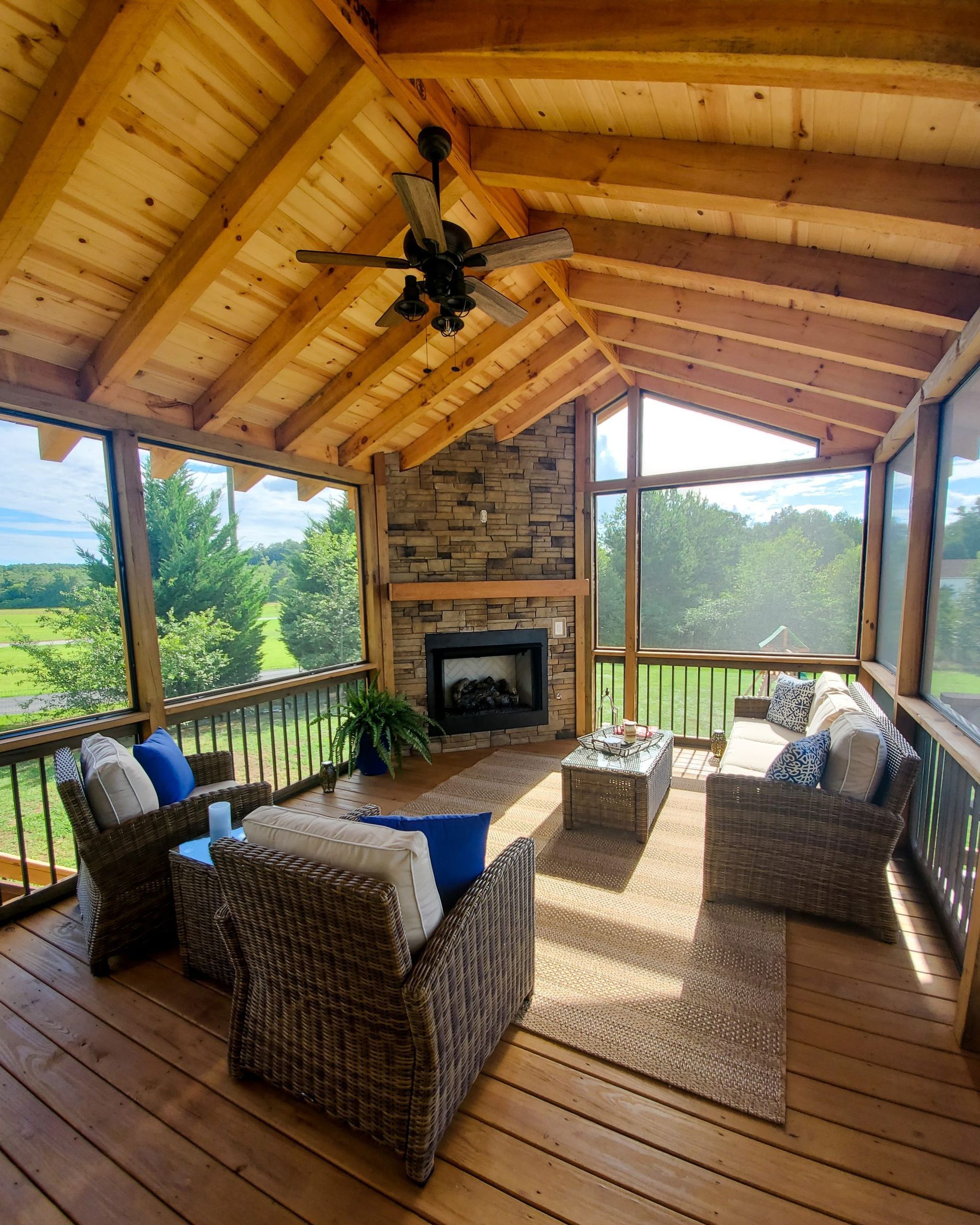 Screened porch with fireplace, wicker furniture, and wood ceiling overlooking a green landscape.