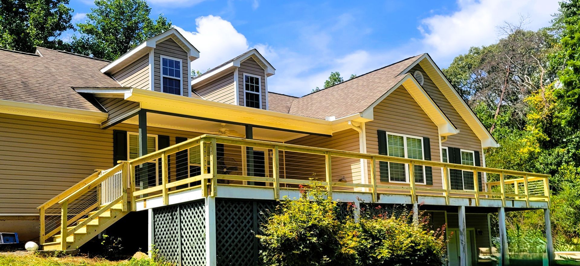 House with yellow porch, deck, and stair railing, surrounded by trees under a blue sky.