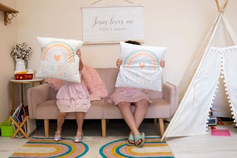 Two girls sitting on a pink couch holding pillows that say you are so loved.