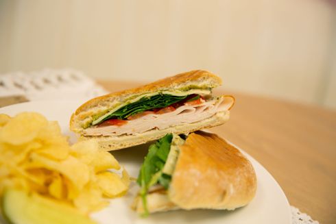 Panini with roasted turkey, spinach, tomato, provolone cheese and basil pesto mayo. Served with chips and a pickle on a white plate at covenant cafe.
