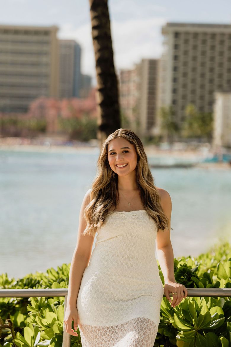 Woman in a white dress standing by the ocean with palm trees and tall buildings in the background