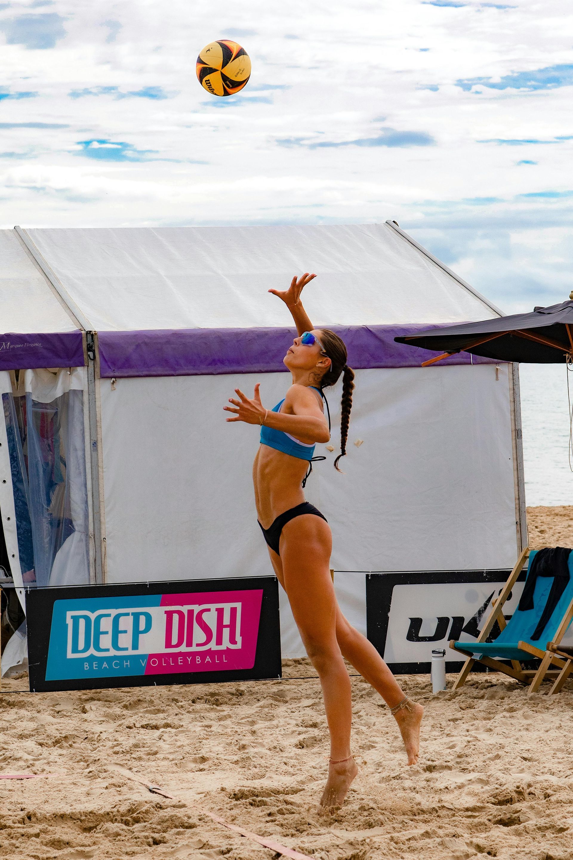 Beach volleyball player in blue bikini reaches up to set a ball on a sandy court.