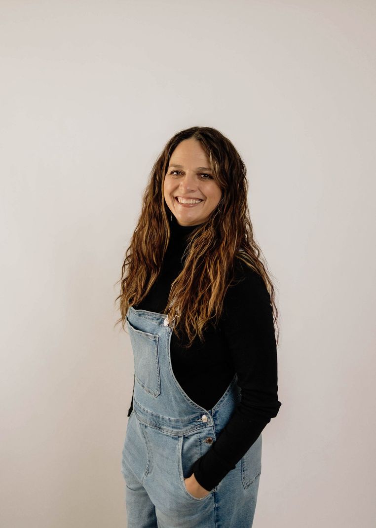 Woman smiling in denim overalls and black top against a plain white wall
