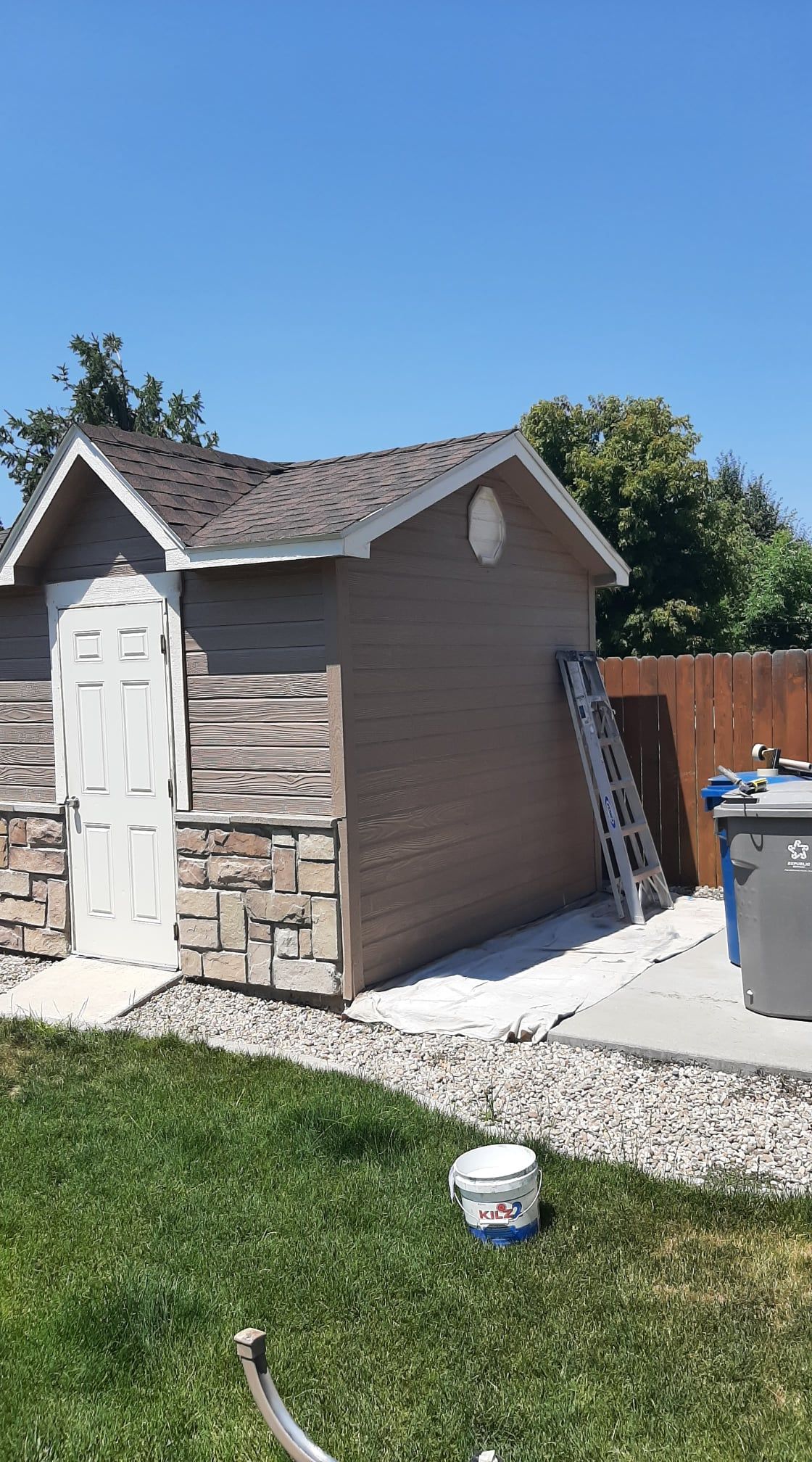 Small brown backyard shed with white doors and stone trim beside a ladder and trash bin on a gravel path.