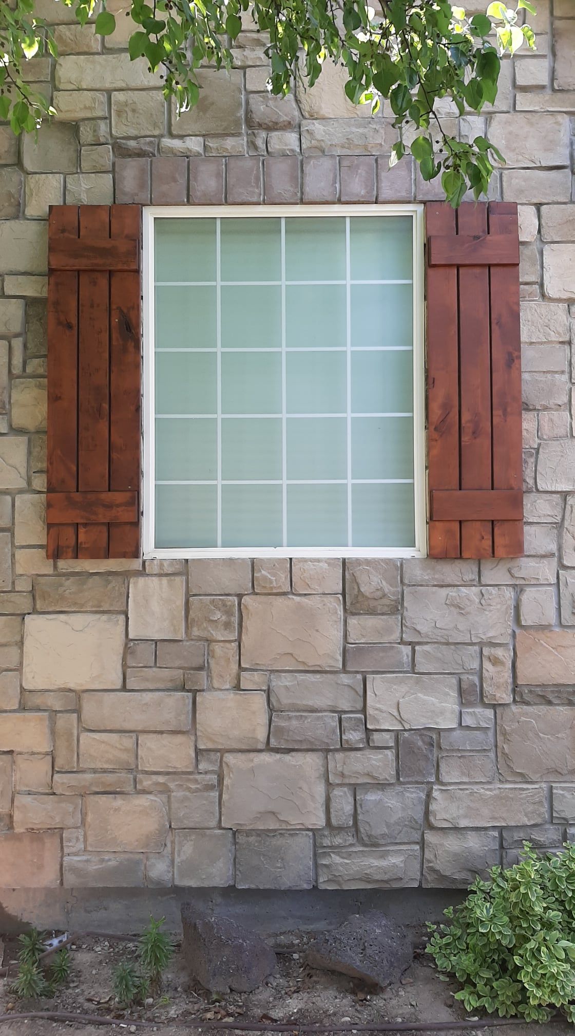 Stone wall with a frosted window framed by red wooden shutters and green leaves above.