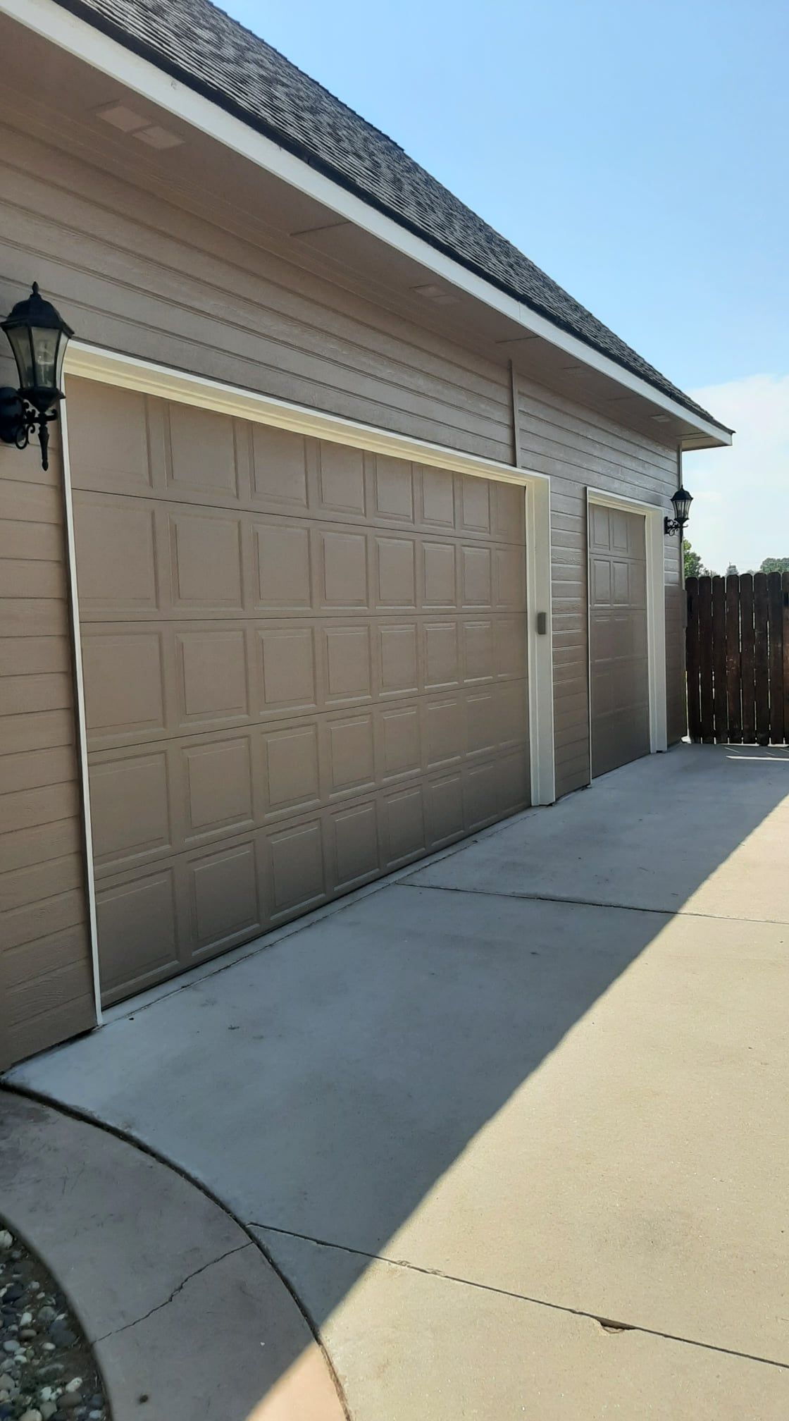 Suburban driveway with beige garage doors, white siding, and a concrete walkway beside a wooden fence
